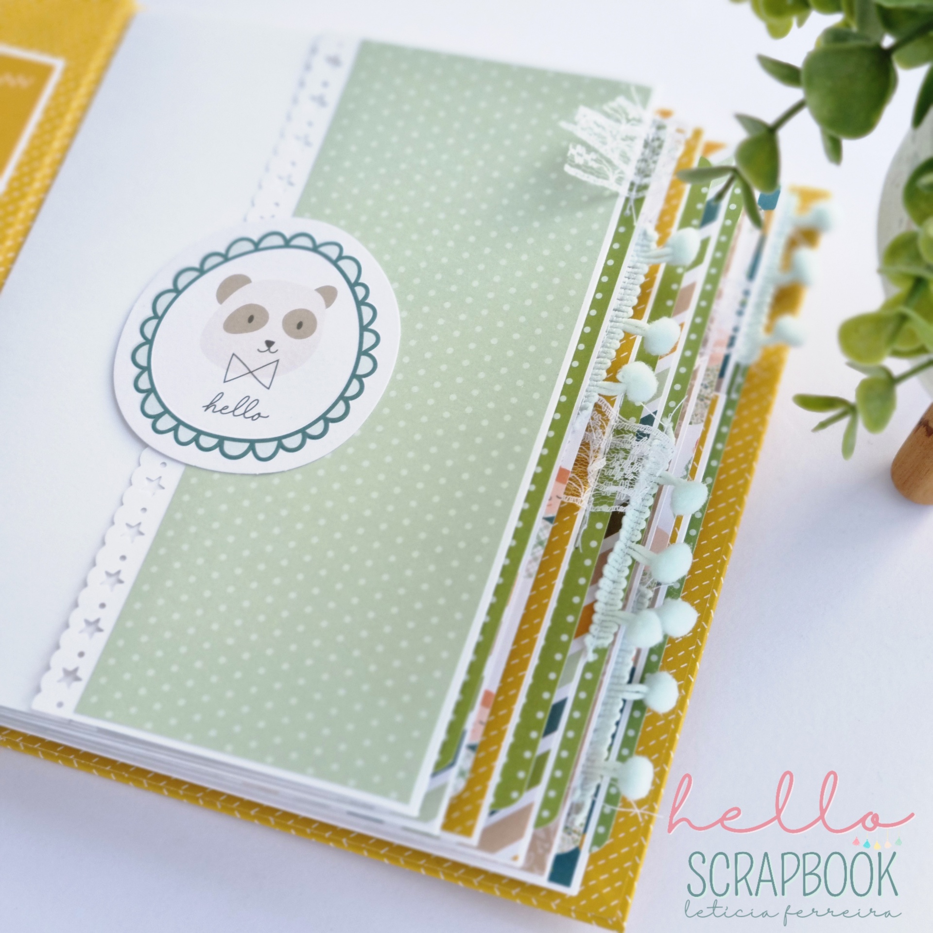 Álbum de scrapbook com capa amarela e páginas decoradas com padrão verde, pompons azuis e etiqueta de urso