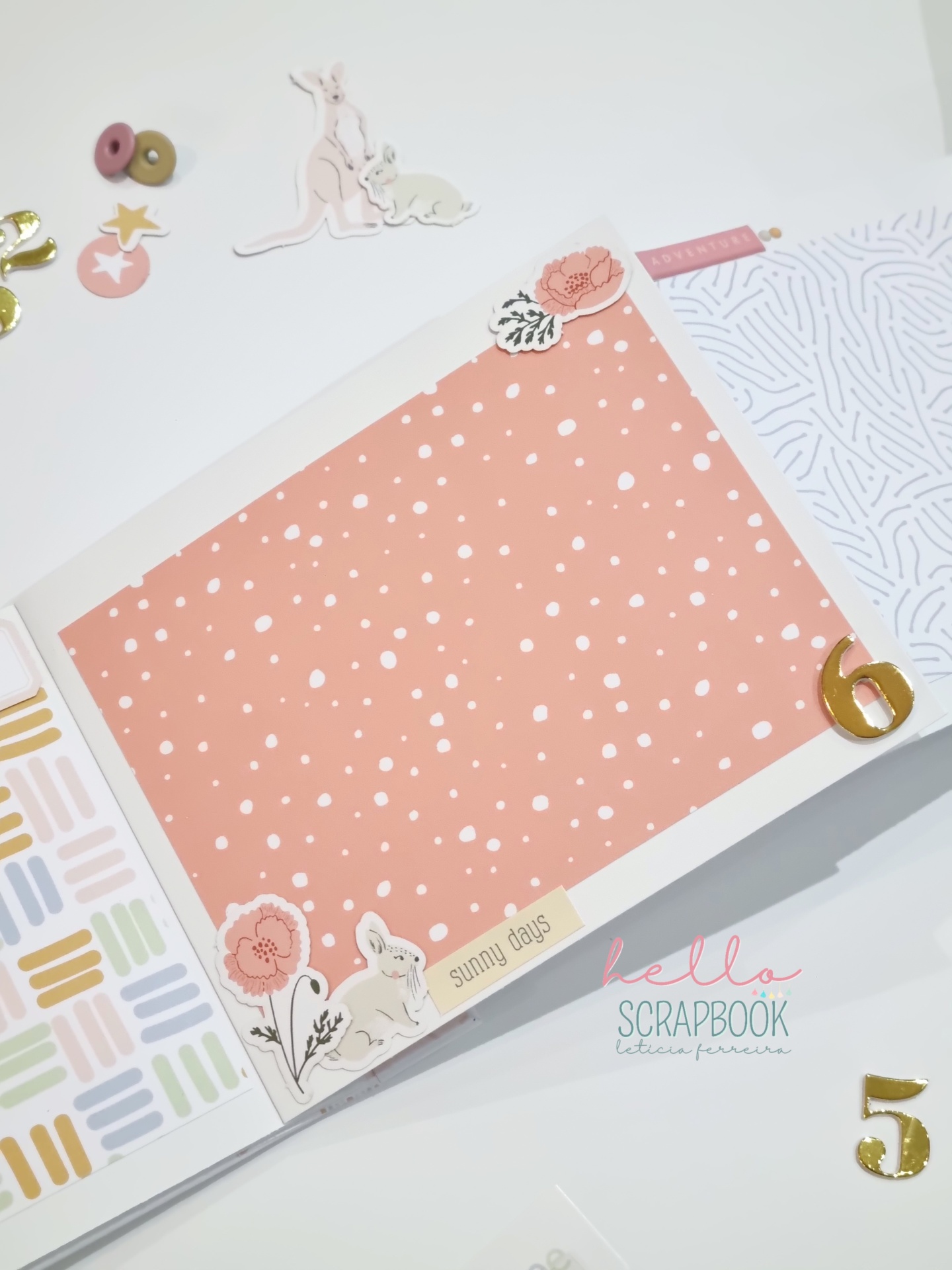 Livro de scrapbook aberto com página central salmão com bolinhas brancas, autocolantes florais e coelho, números dourados 5 e 6 ao redor