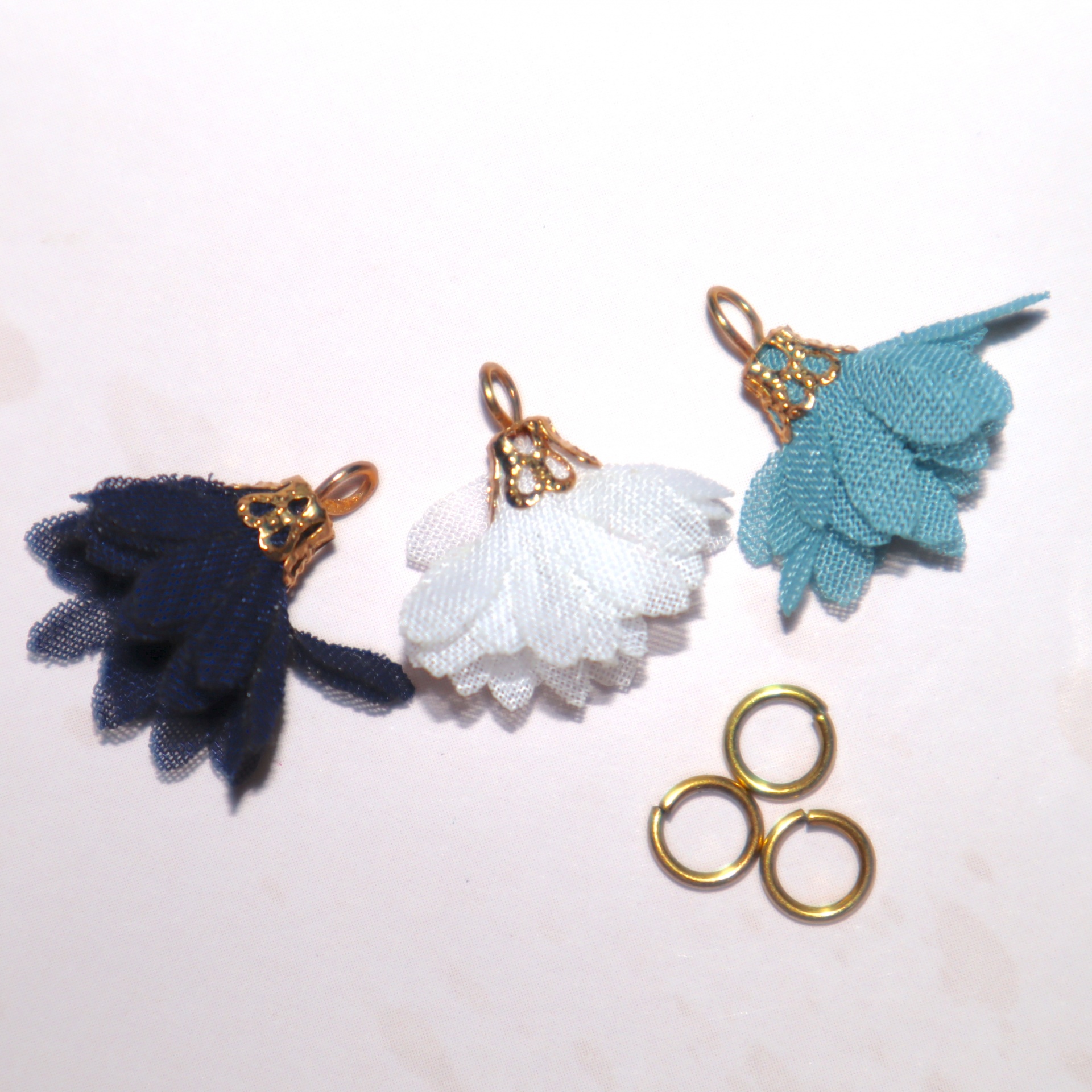 Três pingentes florais de tecido azul escuro, branco e azul claro com detalhes dourados e três argolas douradas sobre fundo branco.