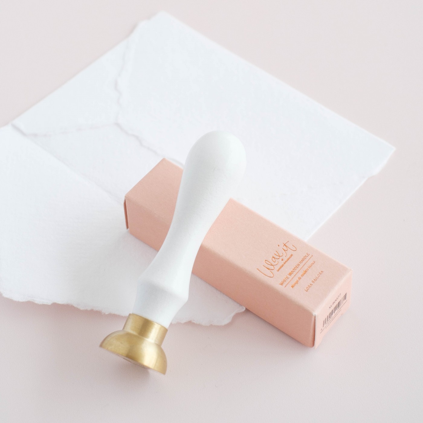 Selo de cera com cabo branco e cabeça dourada e caixa cor de rosa sobre mesa com papéis brancos
