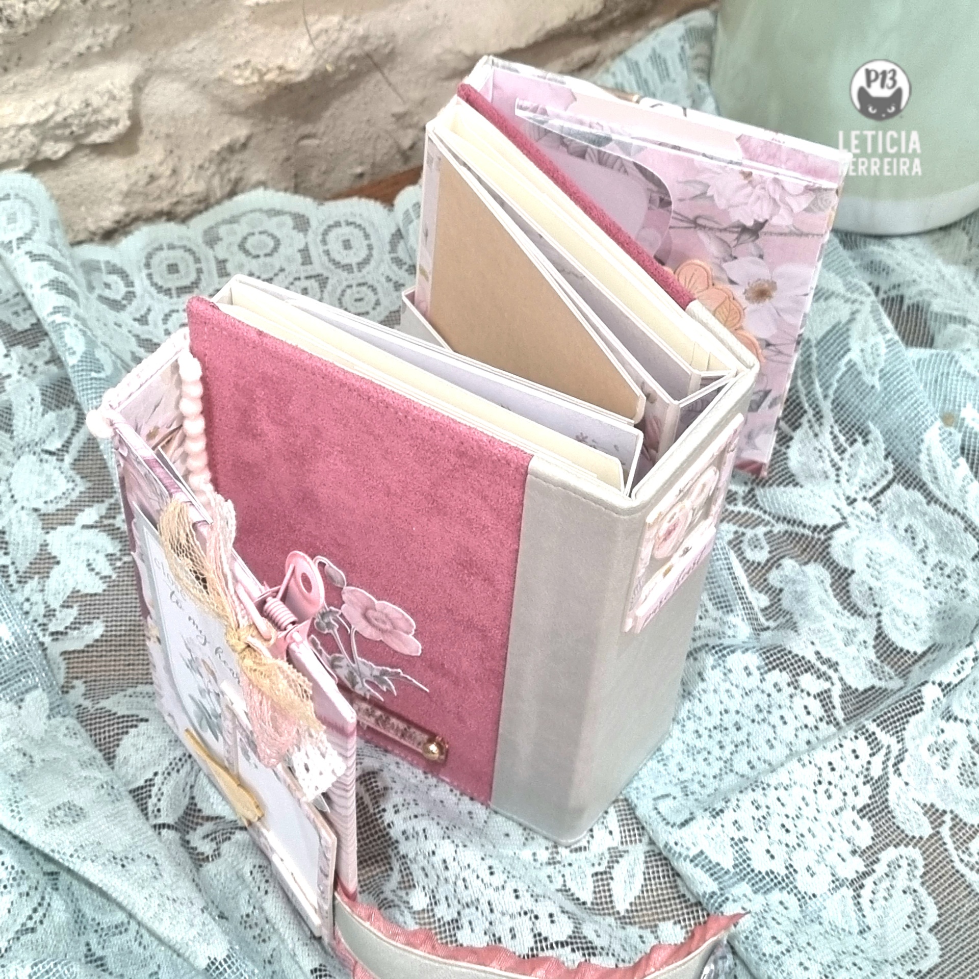 Organizador de tecido rosa e cinza com cadernos e pingentes, sobre toalha branca de renda e parede de pedra