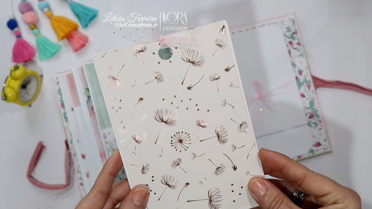 Capa de álbum branco com estampado de sementes de dente-de-leão castanho claro, mãos segurando, fundo com elementos coloridos e caderno aberto