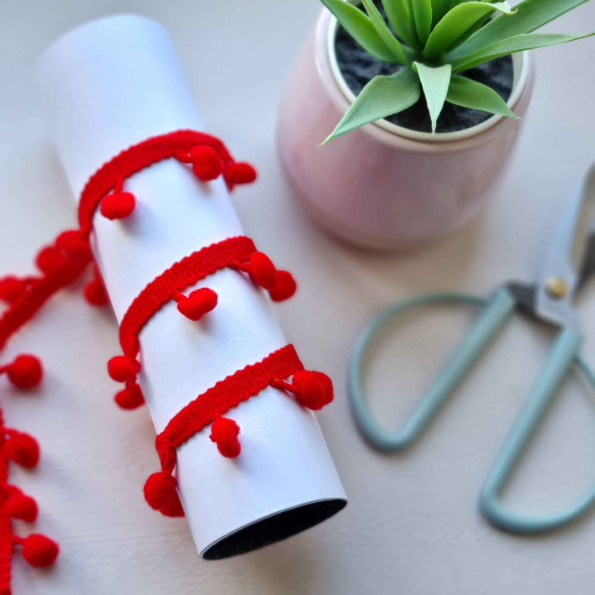 Fita vermelha com pompons enrolada em cilindro branco junto a tesoura e planta em vaso rosa