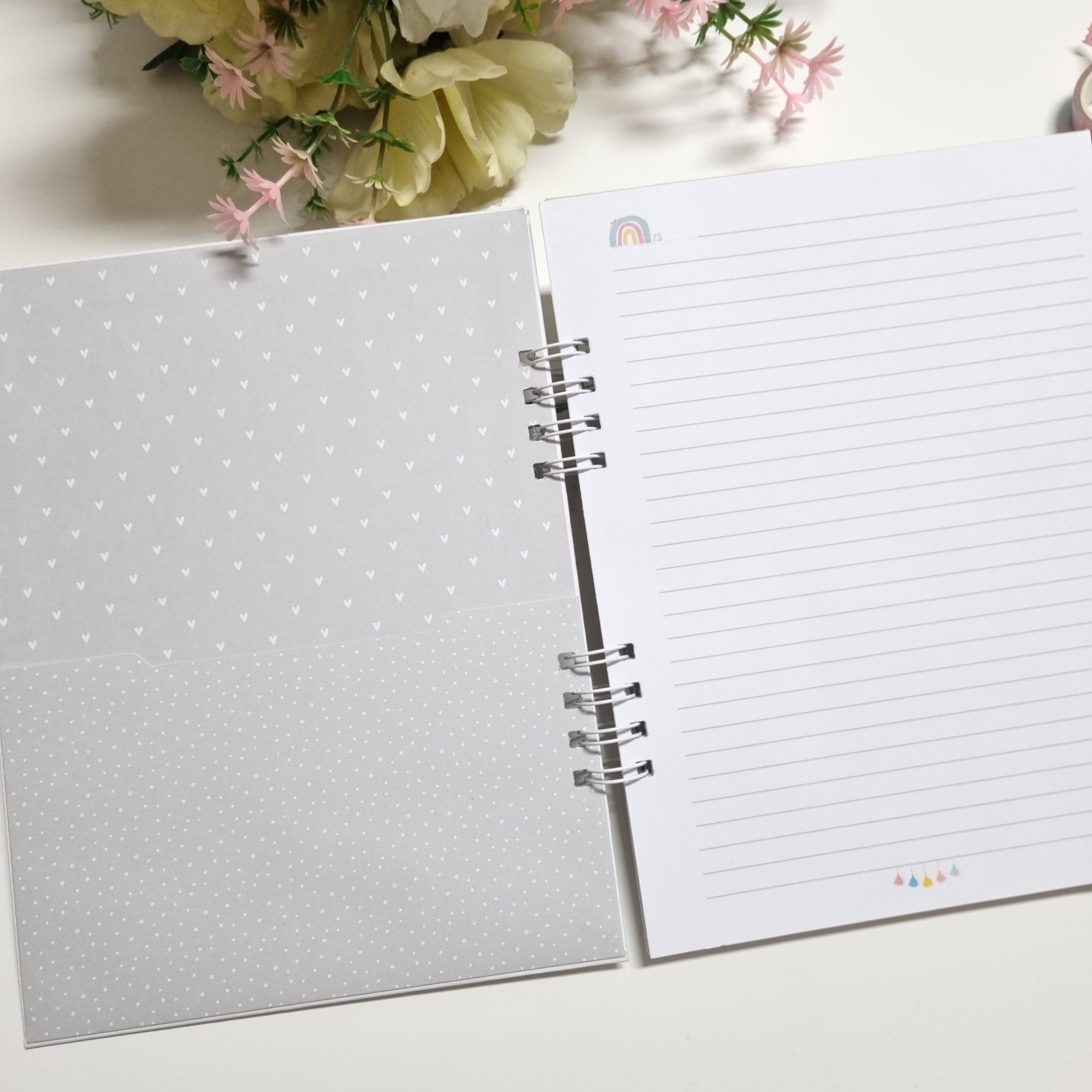 Caderno espiral cinzento com padrão de corações abertos ao lado de flores artificiais