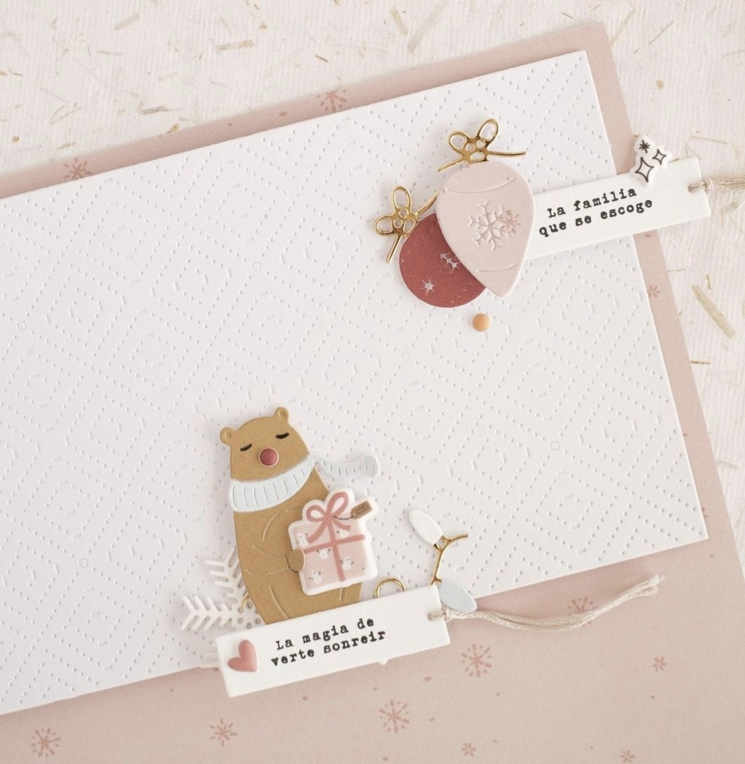 Cartão decorativo branco com urso, rena, presentes e etiquetas com texto em espanhol, sobre fundo bege com flocos de neve.