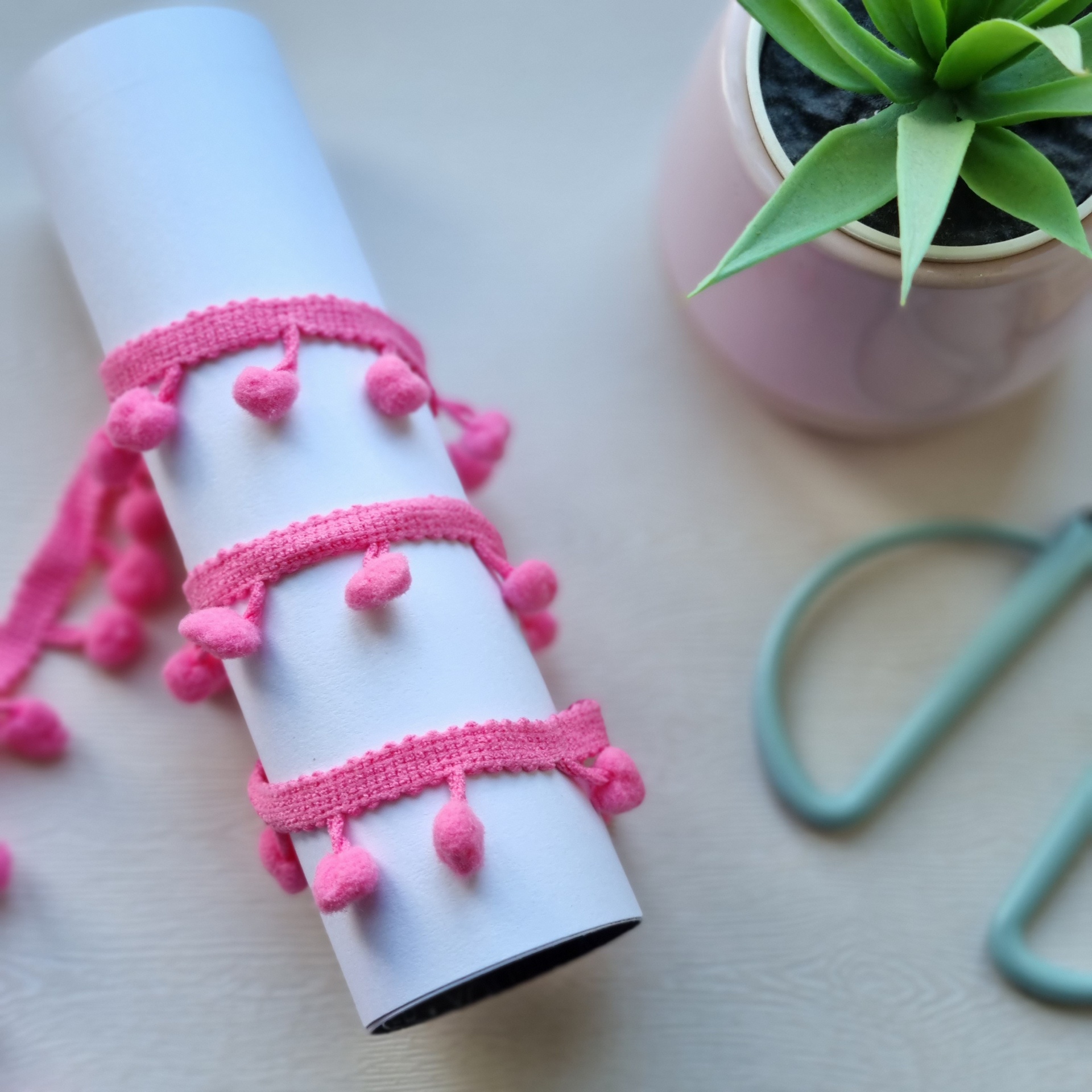 Fitas decorativas cor-de-rosa enroladas num tubo branco junto a planta suculenta num vaso rosa e ferramenta azul