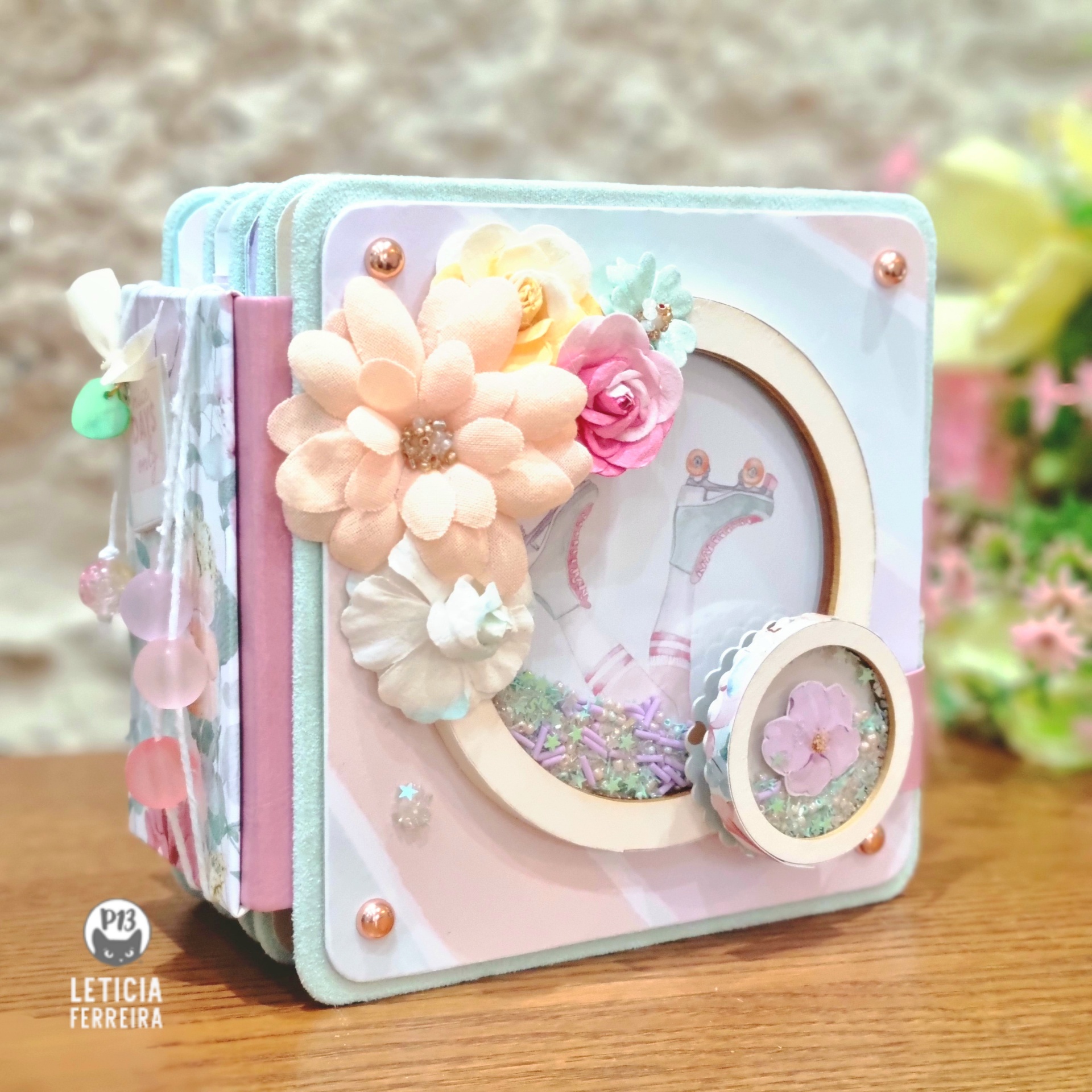 Livro artesanal decorado com flores de tecido em tons pastel e pérolas, sobre mesa de madeira