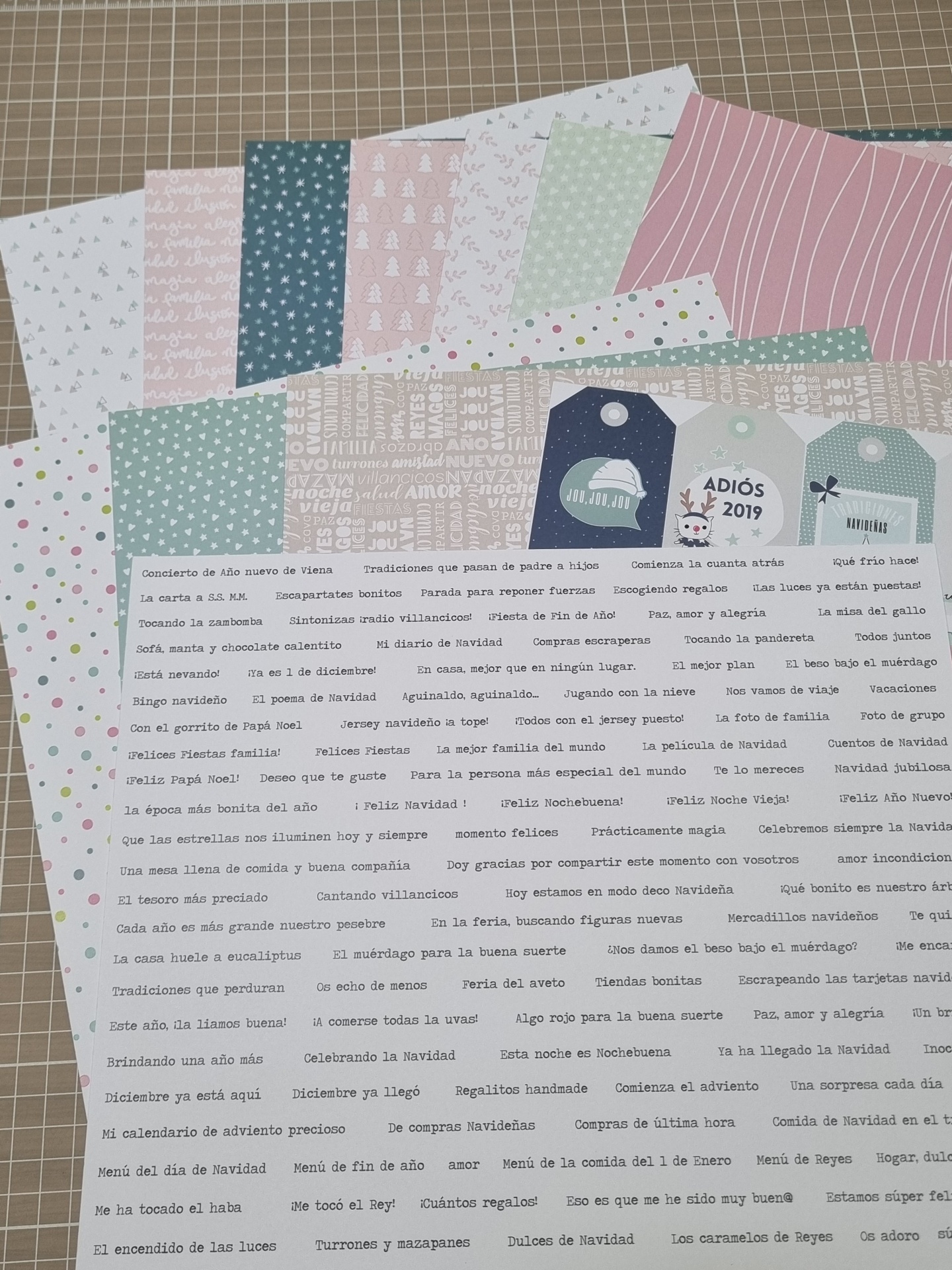 Folhas de papel decorativo com temas natalícios e textos em espanhol sobre uma mesa de hobby com uma superfície de grade.