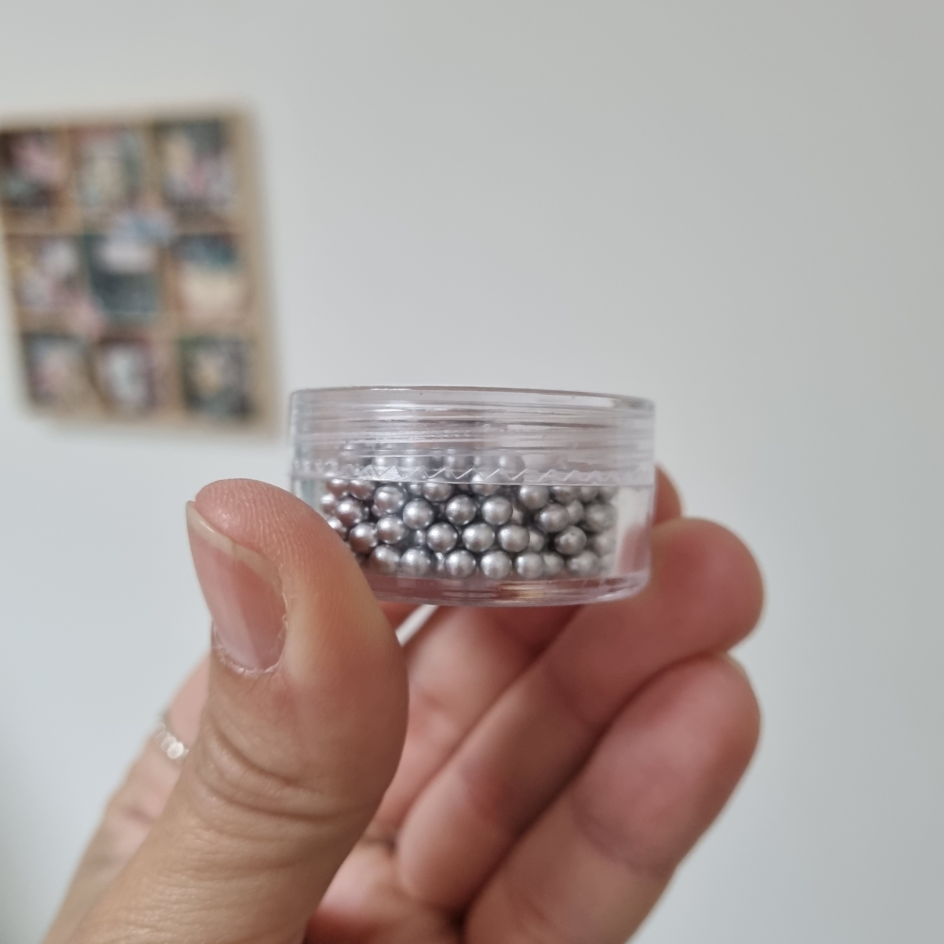 Mão segurando um pequeno recipiente transparente com bolinhas prateadas.