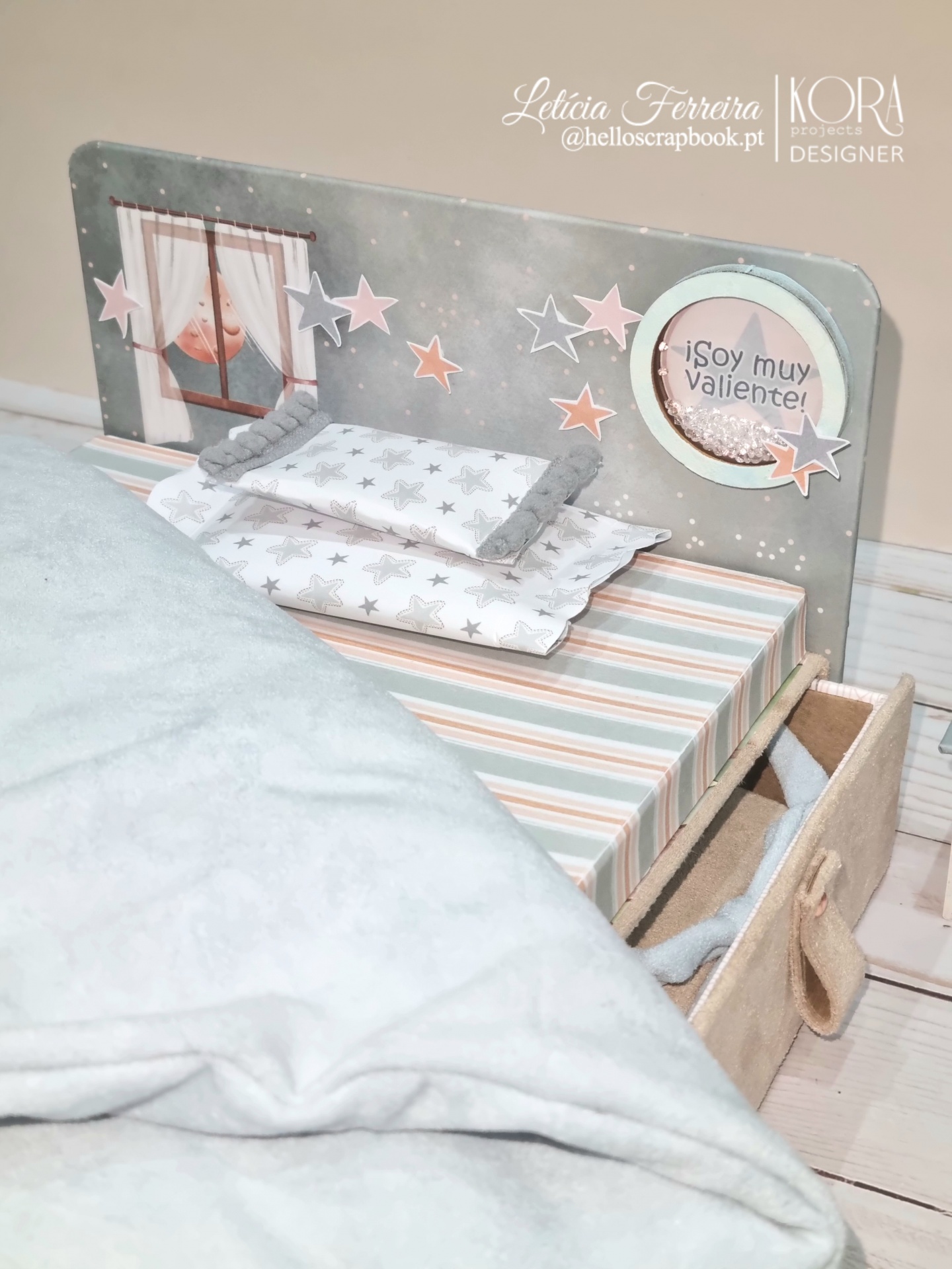 Cama de bebé decorativa com almofadas, manta macia cinzenta e mensagem '¡Soy muy Valiente!' num círculo