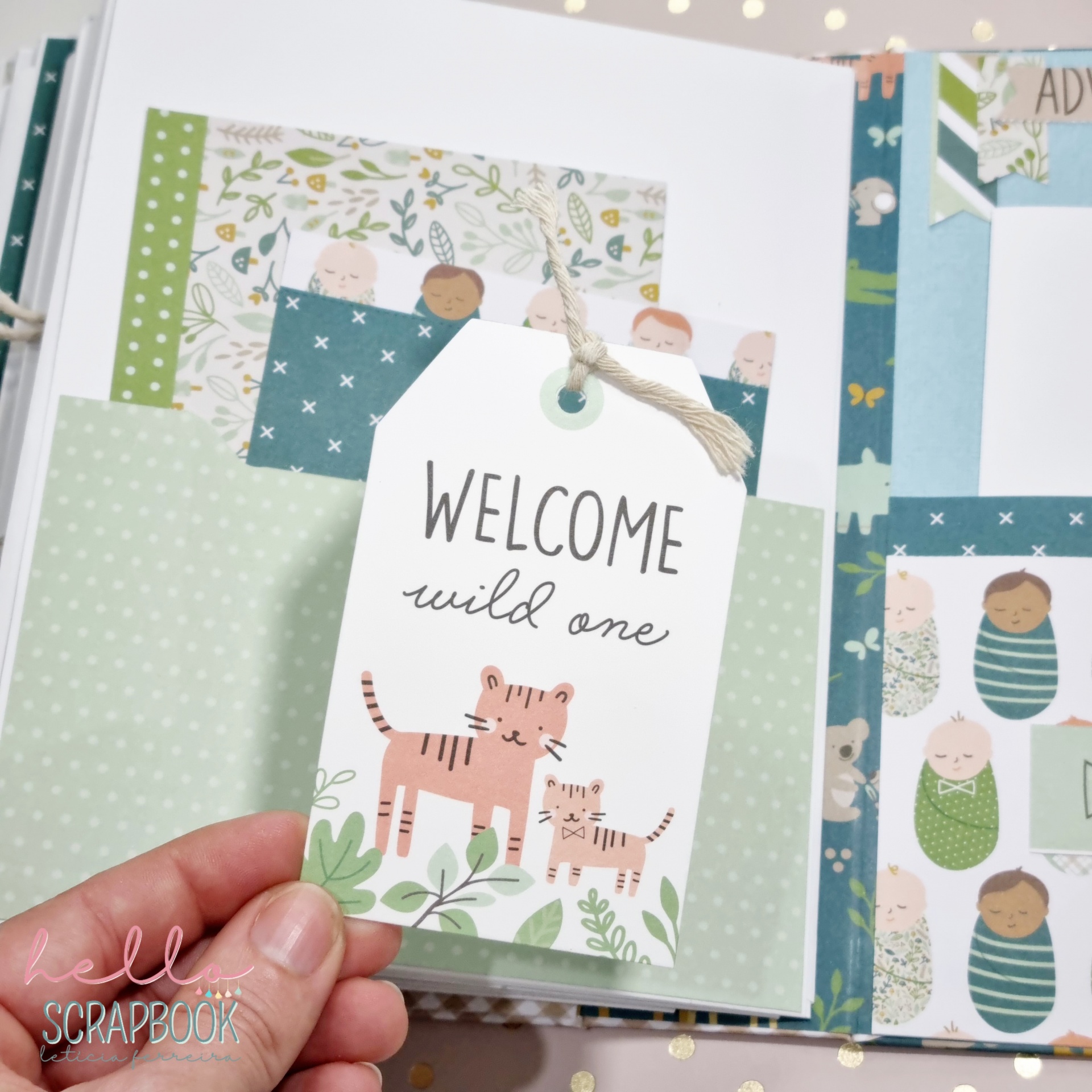 Etiqueta decorativa com tigres e texto 'WELCOME wild one', à frente de álbum de scrapbook com papéis coloridos e padrões infantis.