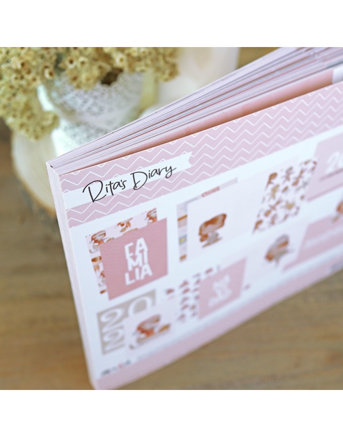 Livro de capa dura rosa com texto 'Rita's Diary' e 'FAMILY' numa mesa de madeira com flores secas ao fundo