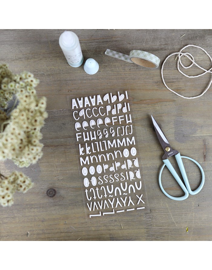 Autocolantes de letras brancas, tesoura azul claro, fio de corda, fita adesiva e frasco branco sobre mesa de madeira.