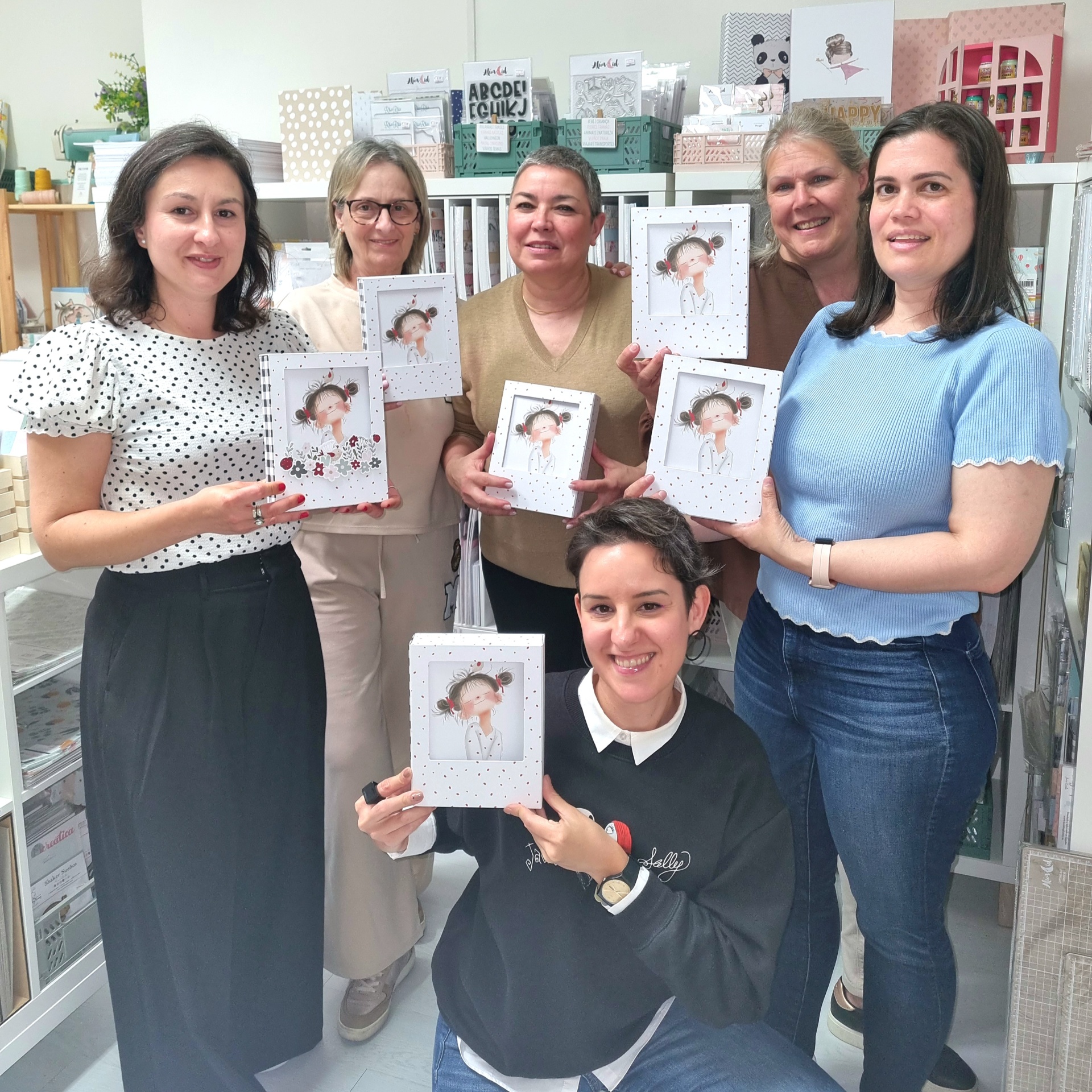 Workshop Menina à Janela Seis mulheres seguram livros com capas brancas e imagem de criança sorridente, num ambiente interior com prateleiras organizadas.