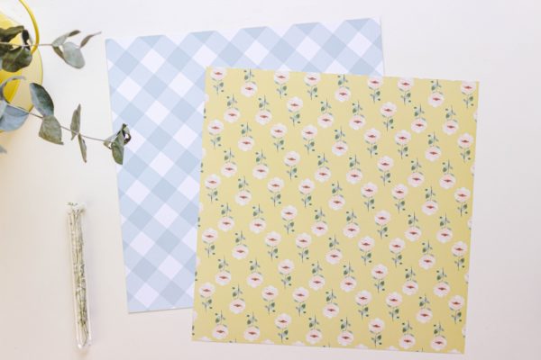 Dois papéis quadrados para artesanato com padrões diferentes, um azul claro xadrez e outro amarelo com flores.