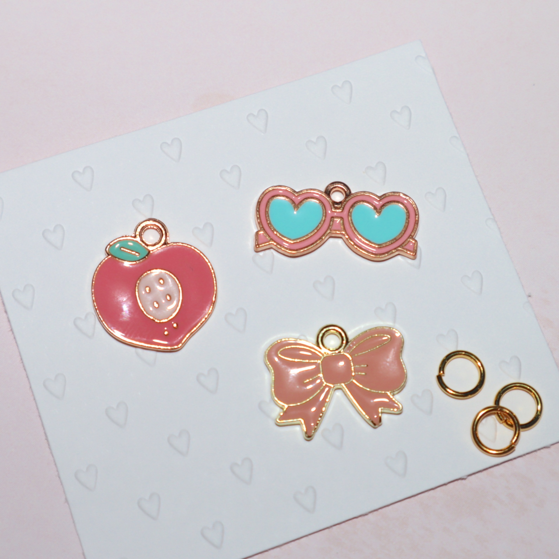 Três pingentes coloridos com acabamento dourado em cartão branco com pequenos corações cinzentos