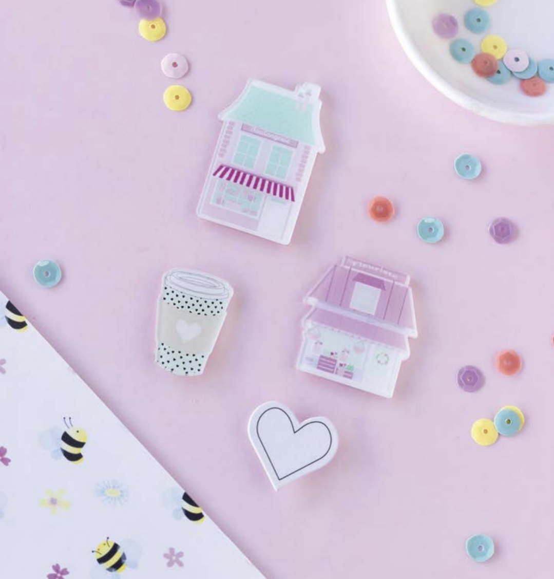 Pins em forma de casas, coração e copo sobre superfície rosa com lantejoulas e tecido com abelhas