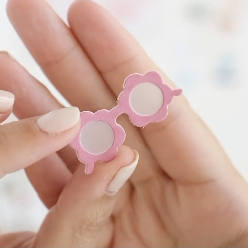 Mini óculos rosa em formato de flor segurados por mãos com unhas brancas
