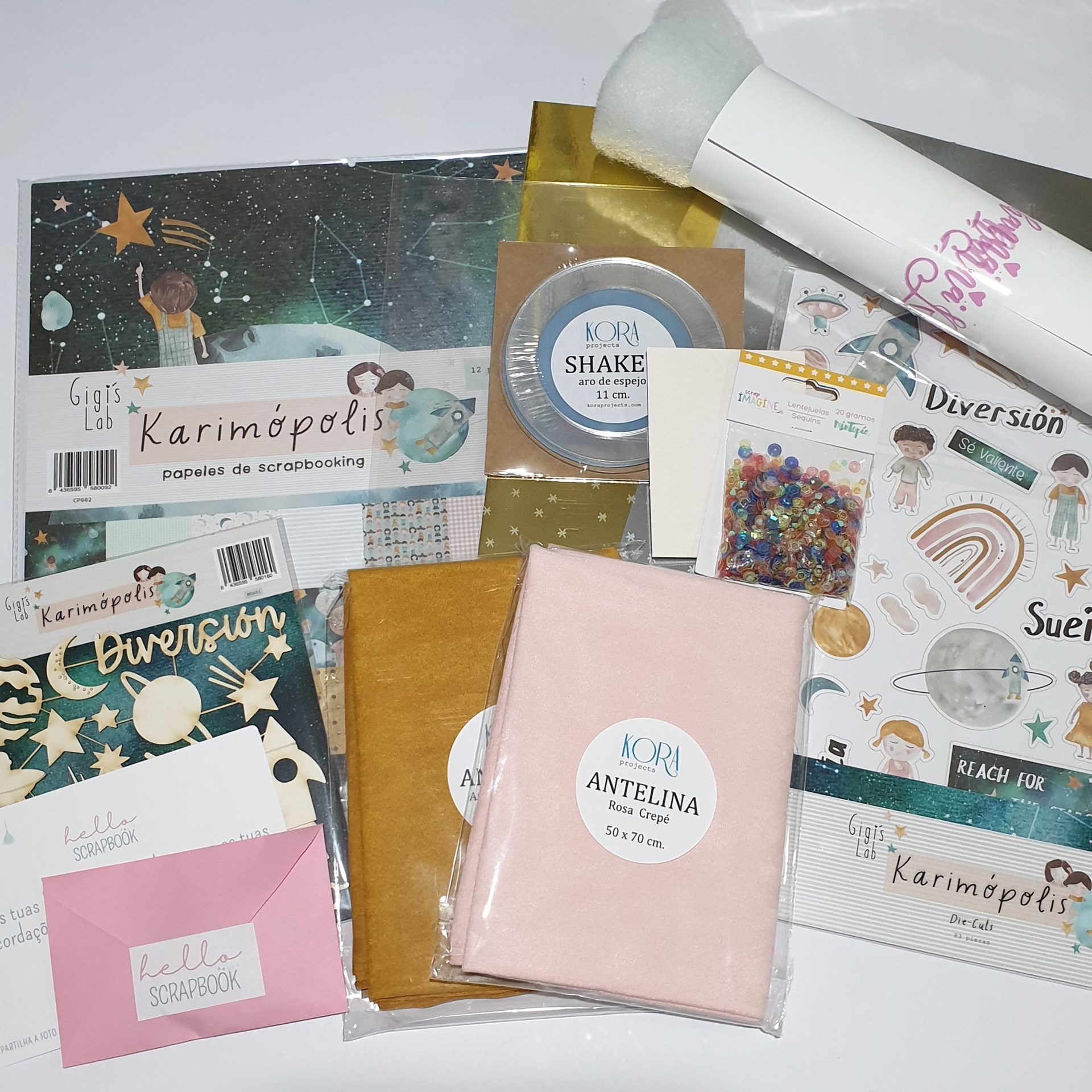 Materiais para scrapbook incluindo papéis decorativos, adesivos, tecidos rosa e mostarda, e pincel branco com texto rosa