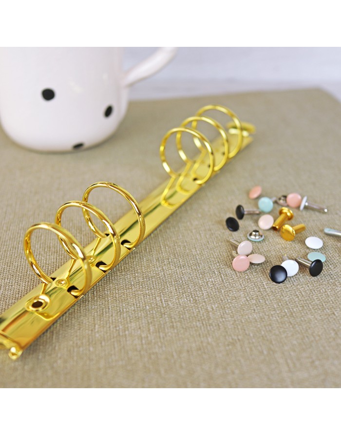 Anel dourado de argolas para fichário com botões coloridos e caneca branca ao fundo.