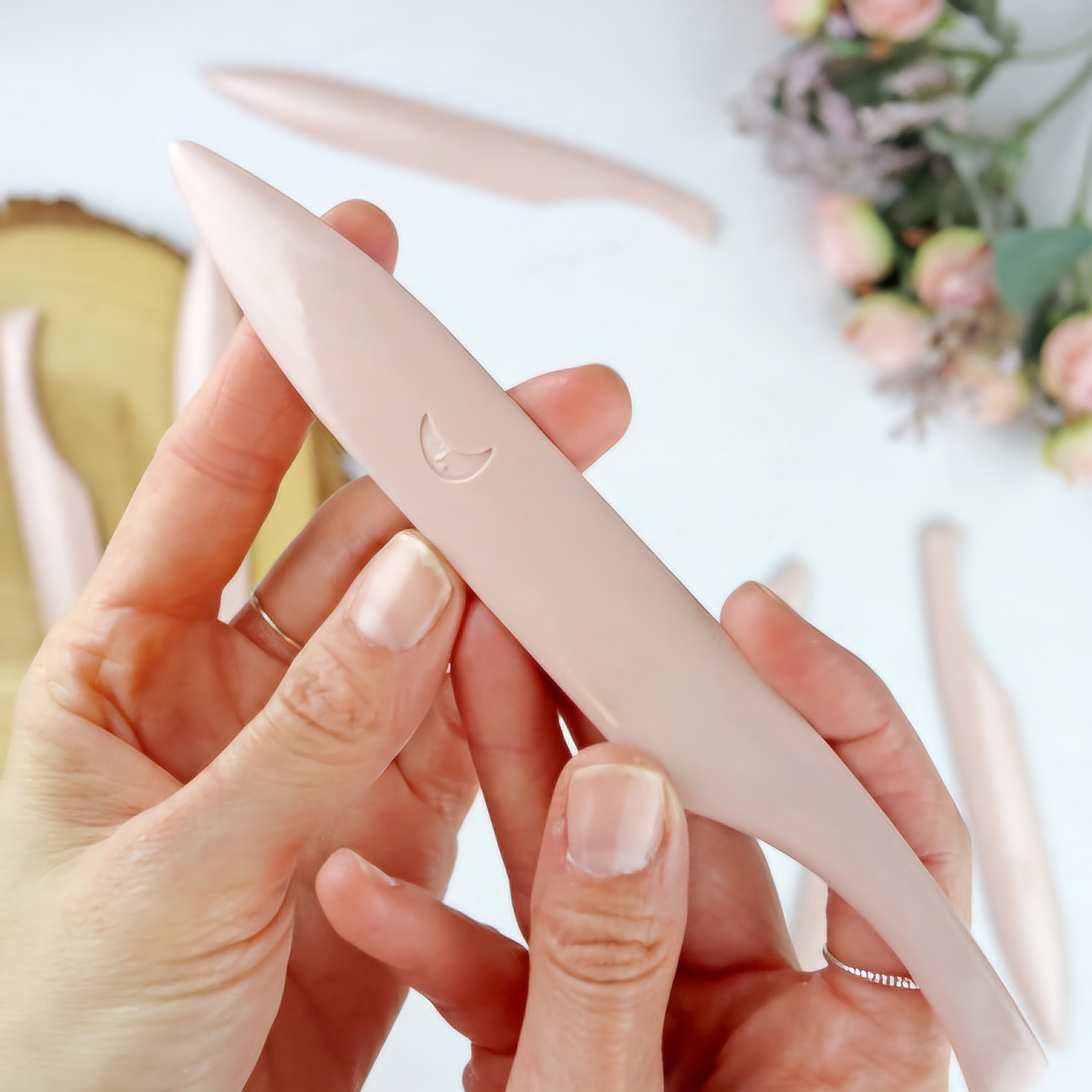Espátula pequena de silicone cor de rosa em mãos, com espátulas e flores ao fundo.