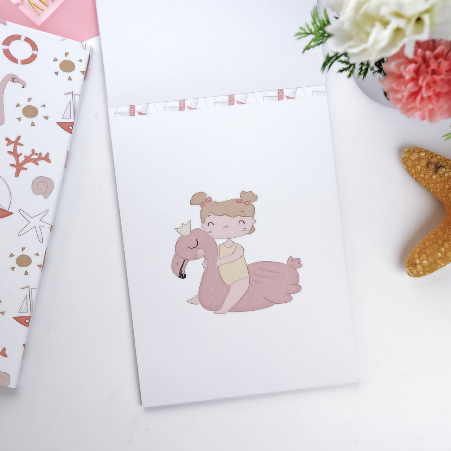 Caderno branco com desenho de rapariga num flamingo rosa e outros objetos decorativos.