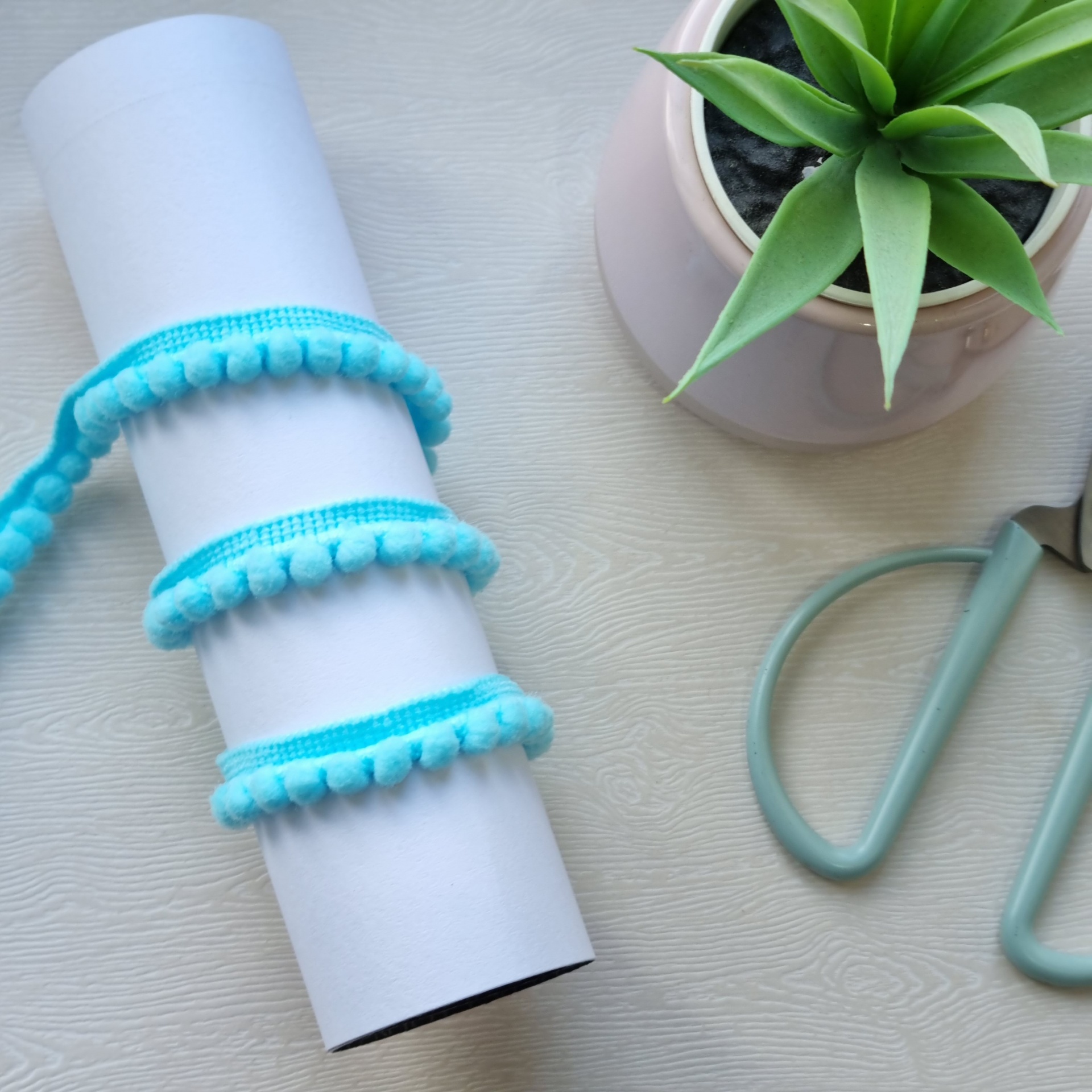 Rolo de tecido com fita azul e planta num vaso branco sobre superfície clara