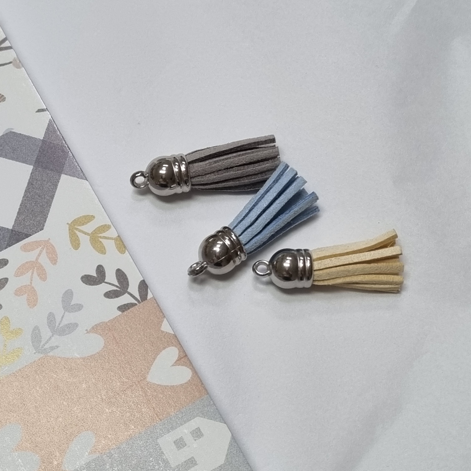 Três borlas de camurça bege, azul claro e cinza com base metálica prateada sobre fundo branco e papel decorativo pastel.