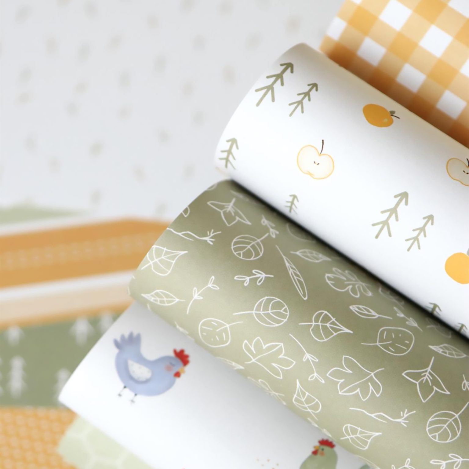 Rolos de papel decorativo com padrões de maçãs, folhas e galinhas sobre fundo branco e verde.