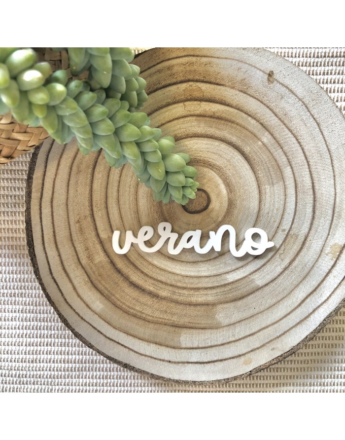 Palavra decorativa 'verano' em madeira branca sobre base circular com textura de madeira e folhas verdes próximas.