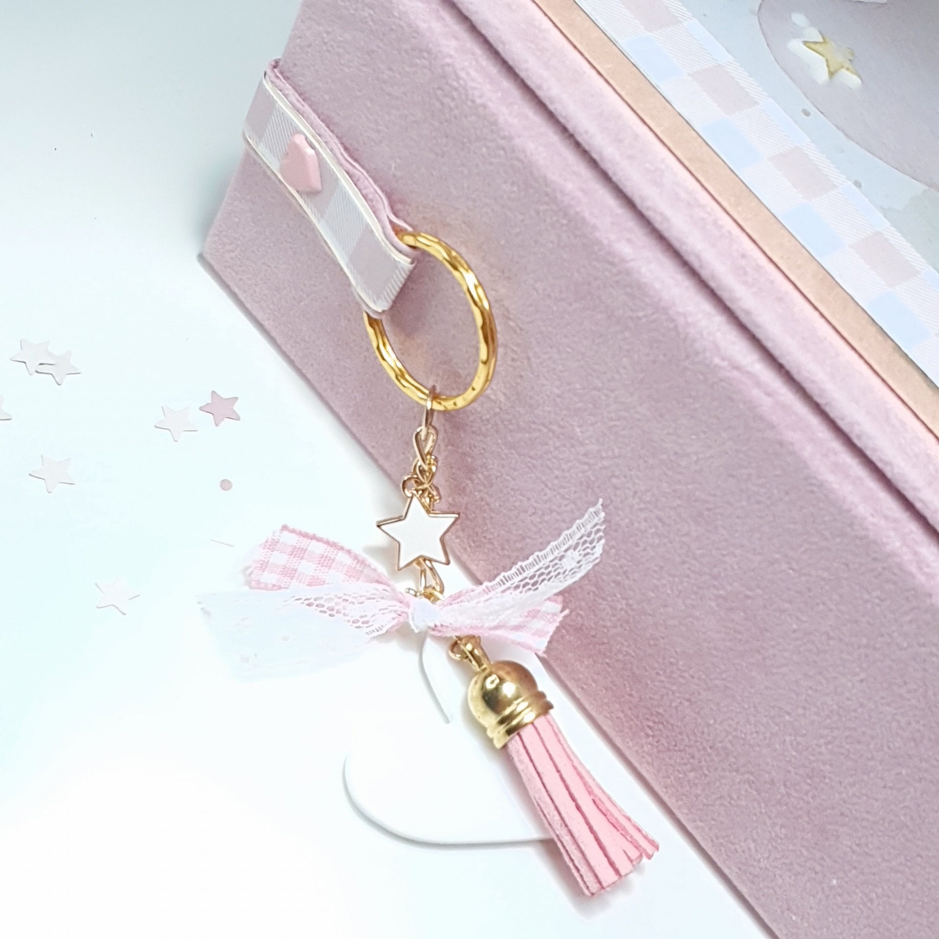 Chaveiro decorativo com argola dourada, estrela branca, fita xadrez rosa e branca, laço de renda branca e borla rosa pendurado em caixa rosada.