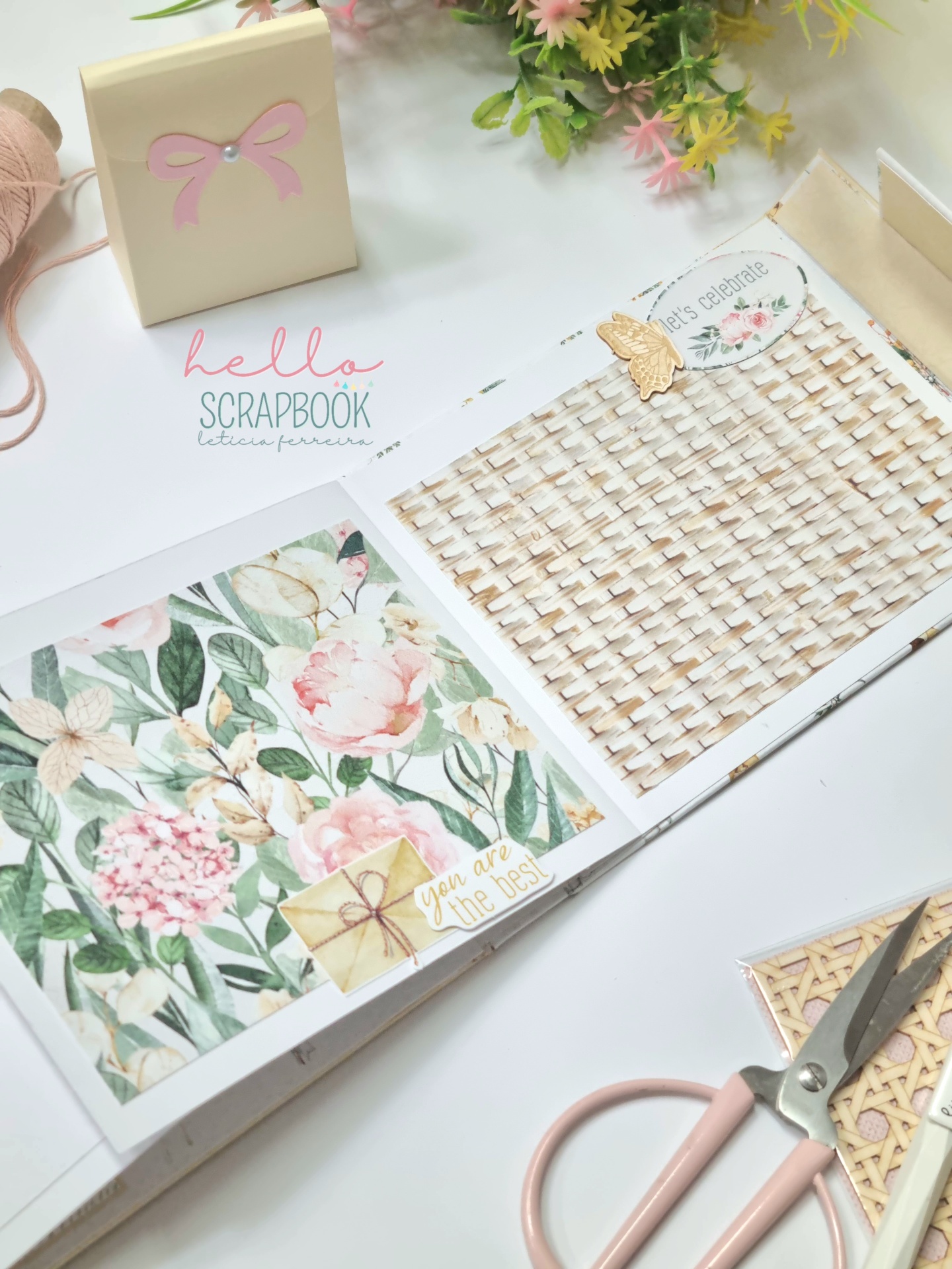Álbum de scrapbooking aberto com padrões florais, tesoura rosa e flores artificiais ao fundo