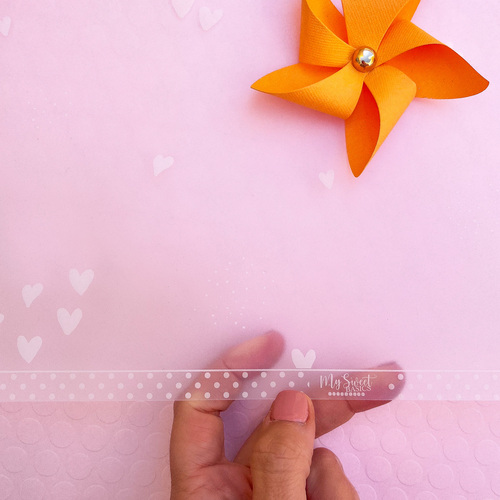 Folha plástica decorativa transparente com corações brancos, sustentada por mão em fundo rosa e moinho laranja.
