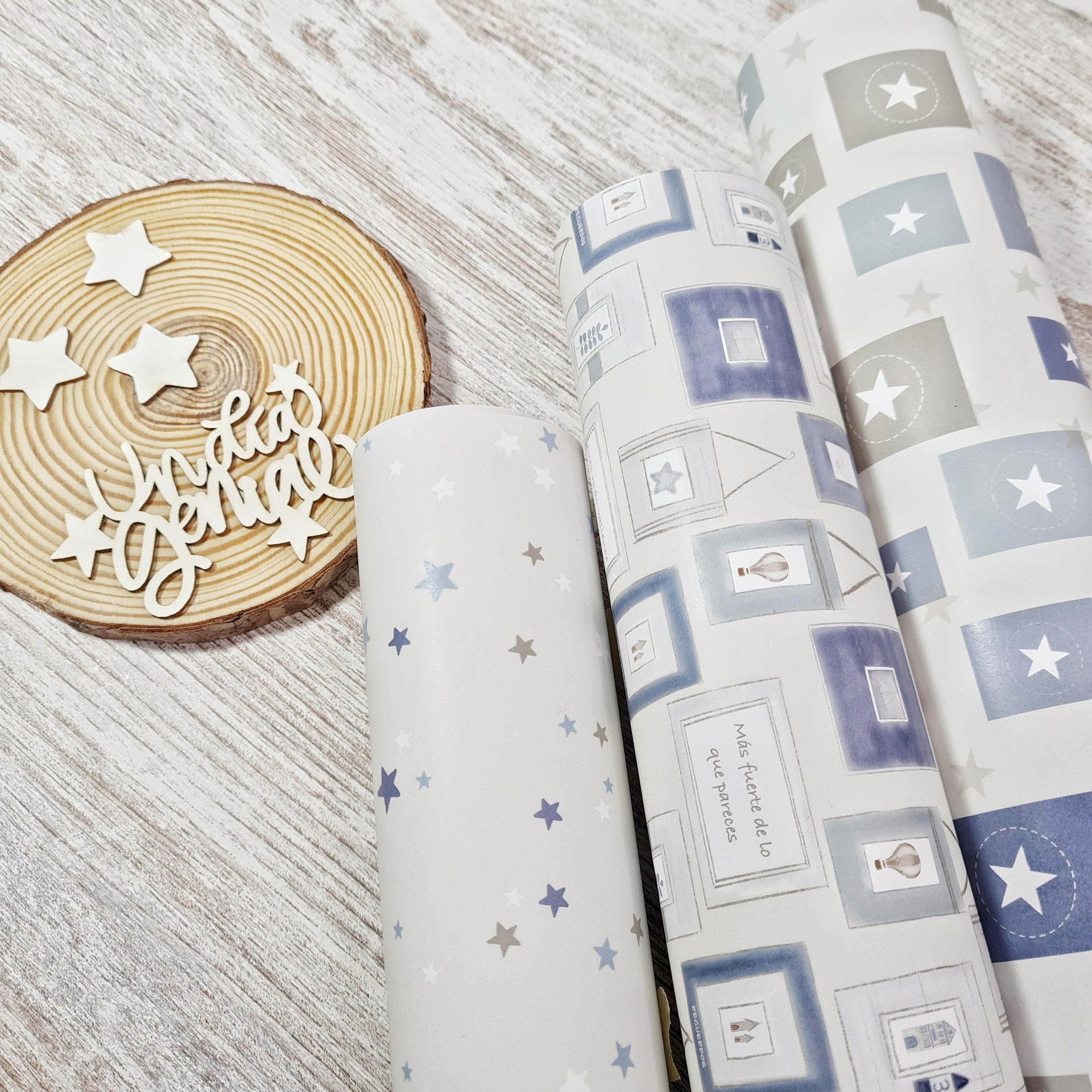 Três rolos de papel de parede com padrões de estrelas e quadrados em azul, cinza e branco sobre madeira clara e fatia de madeira decorativa com estrelas e texto 'muito gelado'