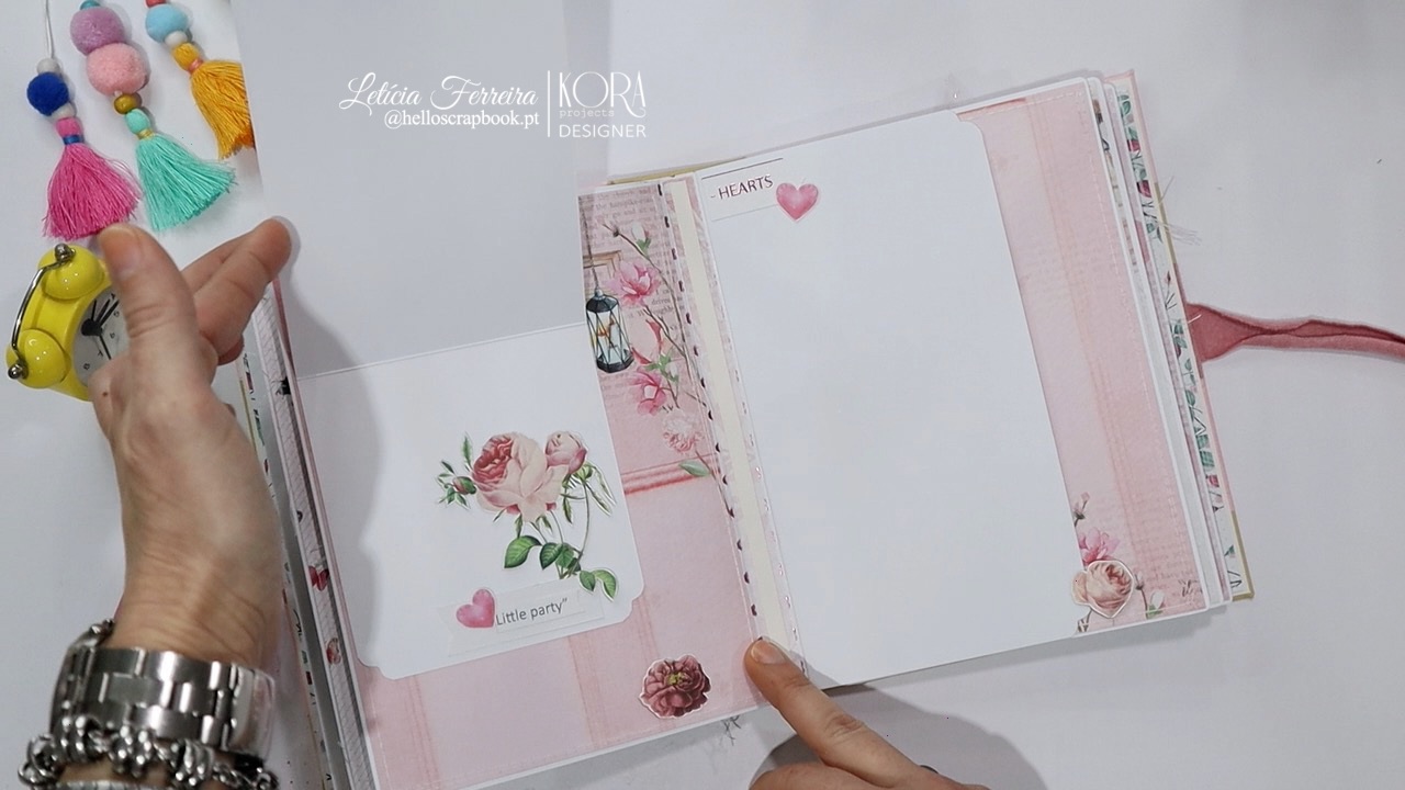 Caderno rosa aberto com decoração floral, cartão com rosas e pompons coloridos ao lado