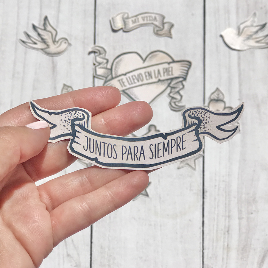 Autocolante em forma de faixa com texto 'JUNTOS PARA SIEMPRE' segurado por mão sobre madeira clara com outros autocolantes. 