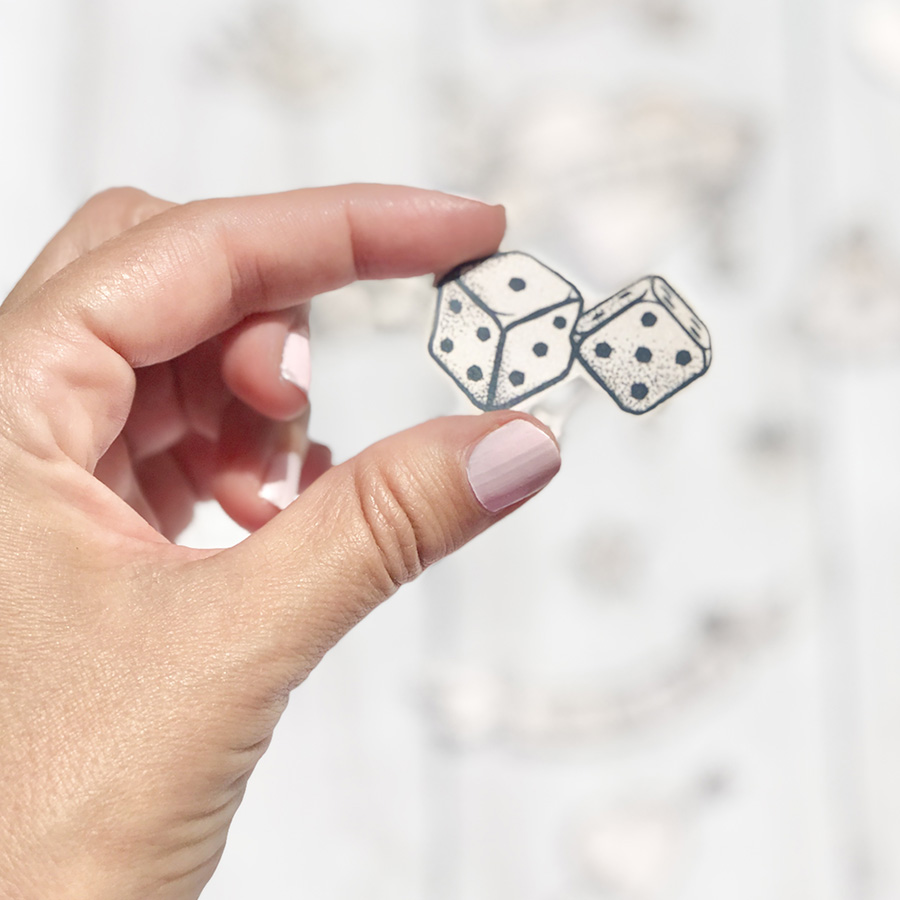 Brincos em forma de dados brancos com pontos pretos sendo segurados por uma mão com unhas rosas claras