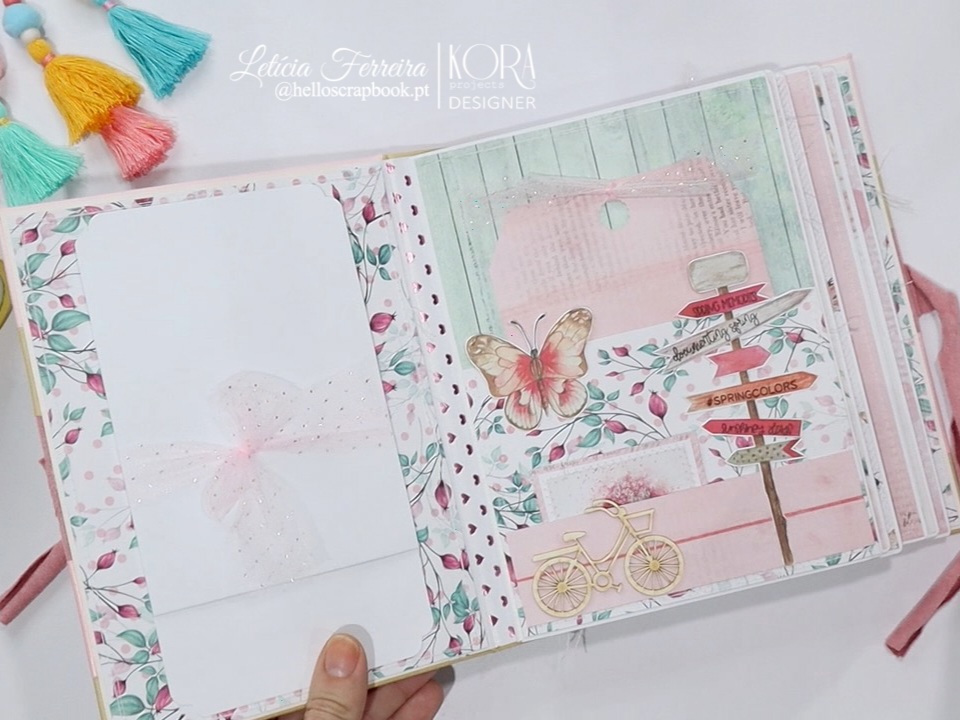 Álbum de scrapbook decorado com padrões florais, bicicleta amarela, borboleta, marcador colorido e envelope branco com laço.