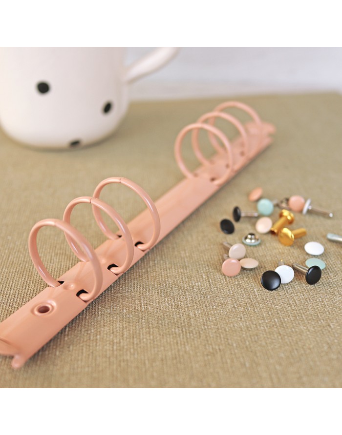 Anel para fichário rosa com argolas e rebites coloridos sobre tecido bege.