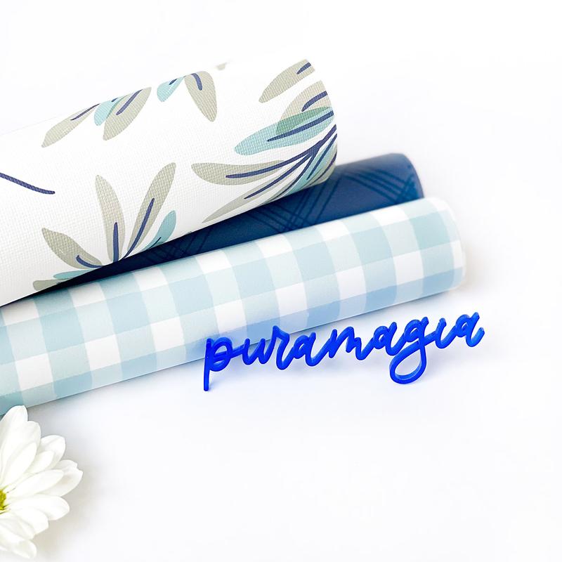 Rolos de papel ou tecido com padrões variados sobre fundo branco, flor branca no canto e texto azul 'puramagia'