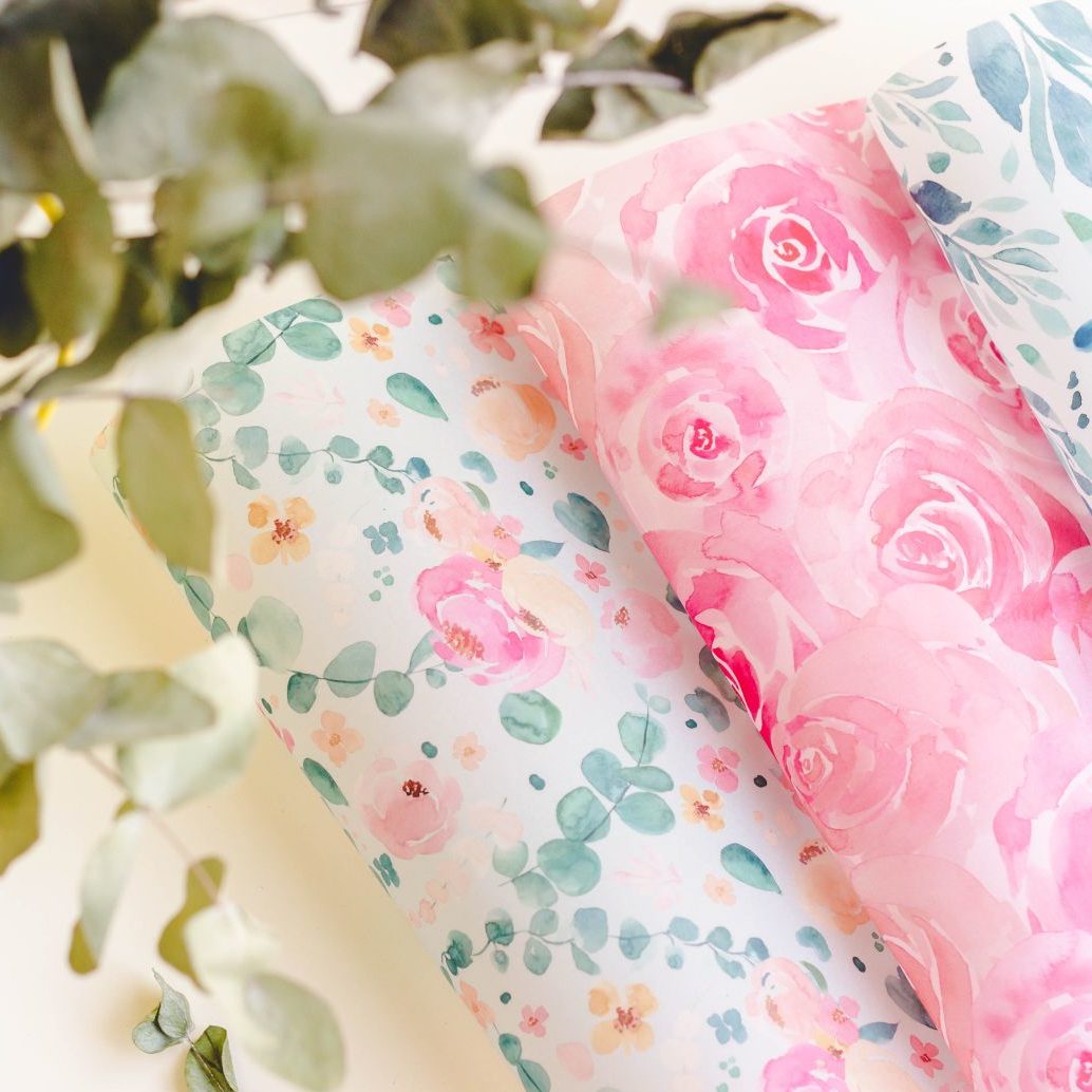 Rolos de papel de embrulho com padrões florais em tons de rosa, azul e verde com folhas desfocadas em primeiro plano