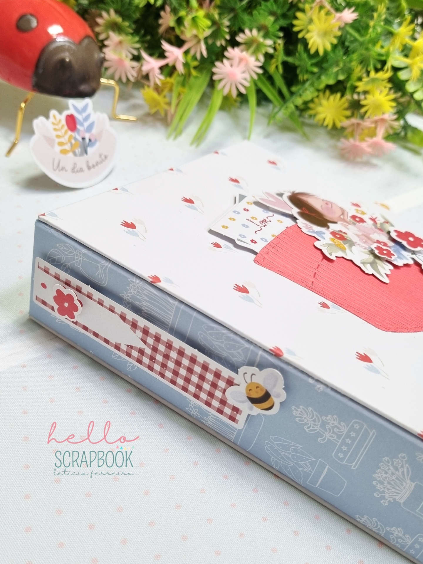 Caixa de scrapbook decorada com padrões florais e abelha com flores artificiais ao fundo