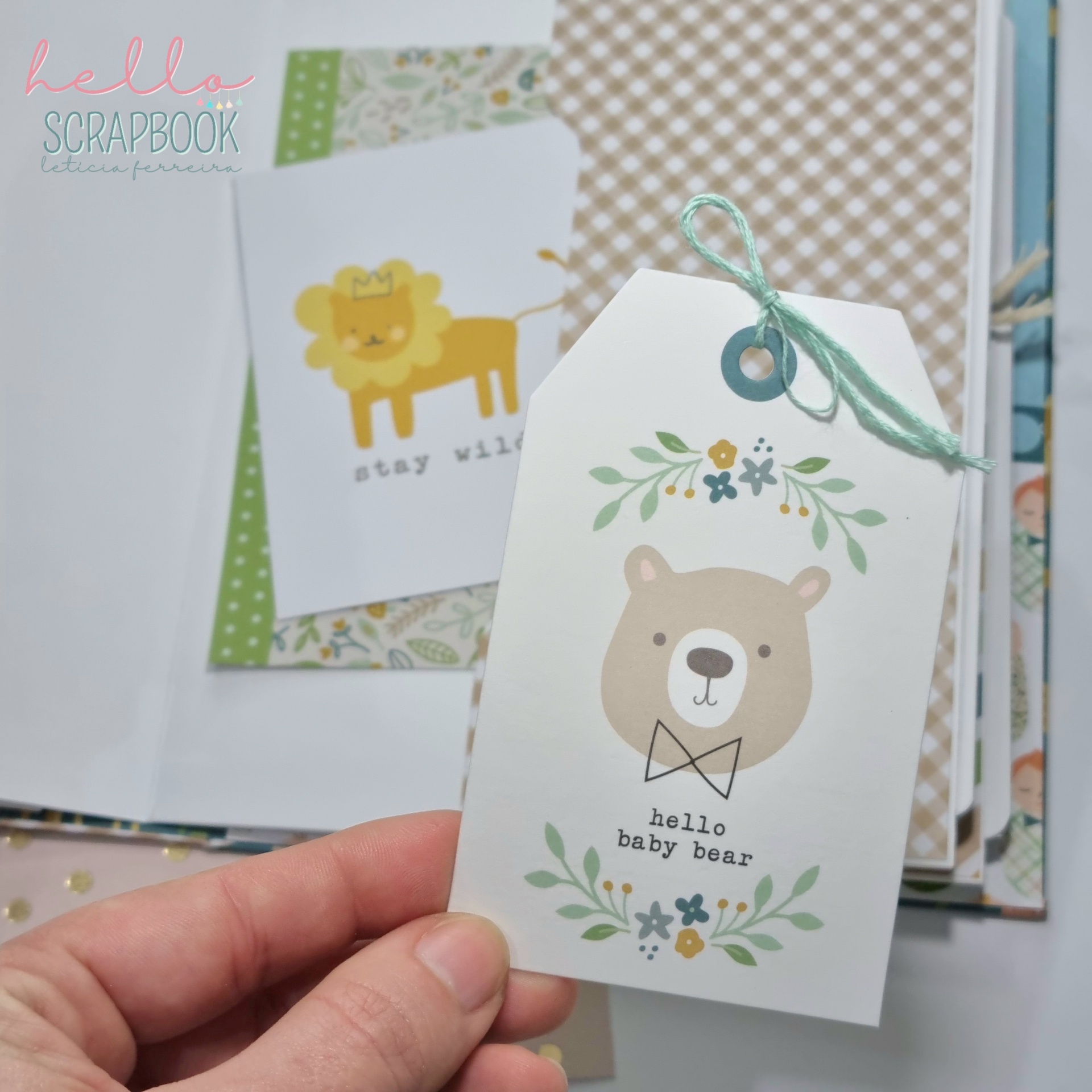 Etiqueta branca com ilustração de urso e texto 'hello baby bear', segurada por mão, com scrapbook ao fundo
