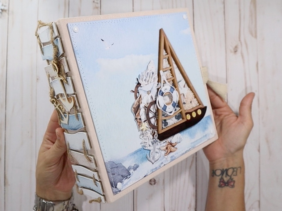 Álbum scrapbook com capa azul e veleiro de madeira, conchas e cordas, mãos segurando