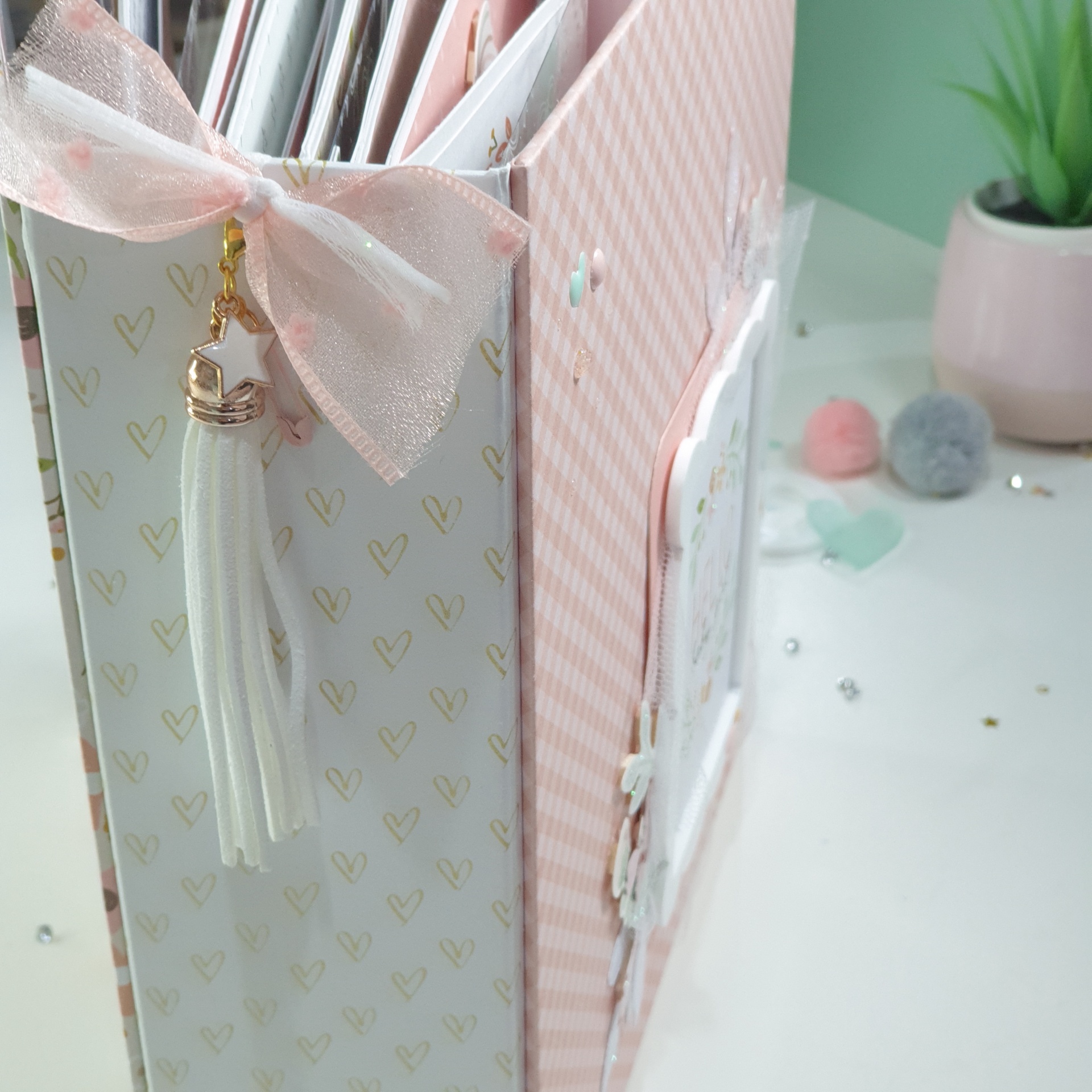 Organizador de papelaria com padrão de corações dourados, laço e tassel branco em mesa decorada.