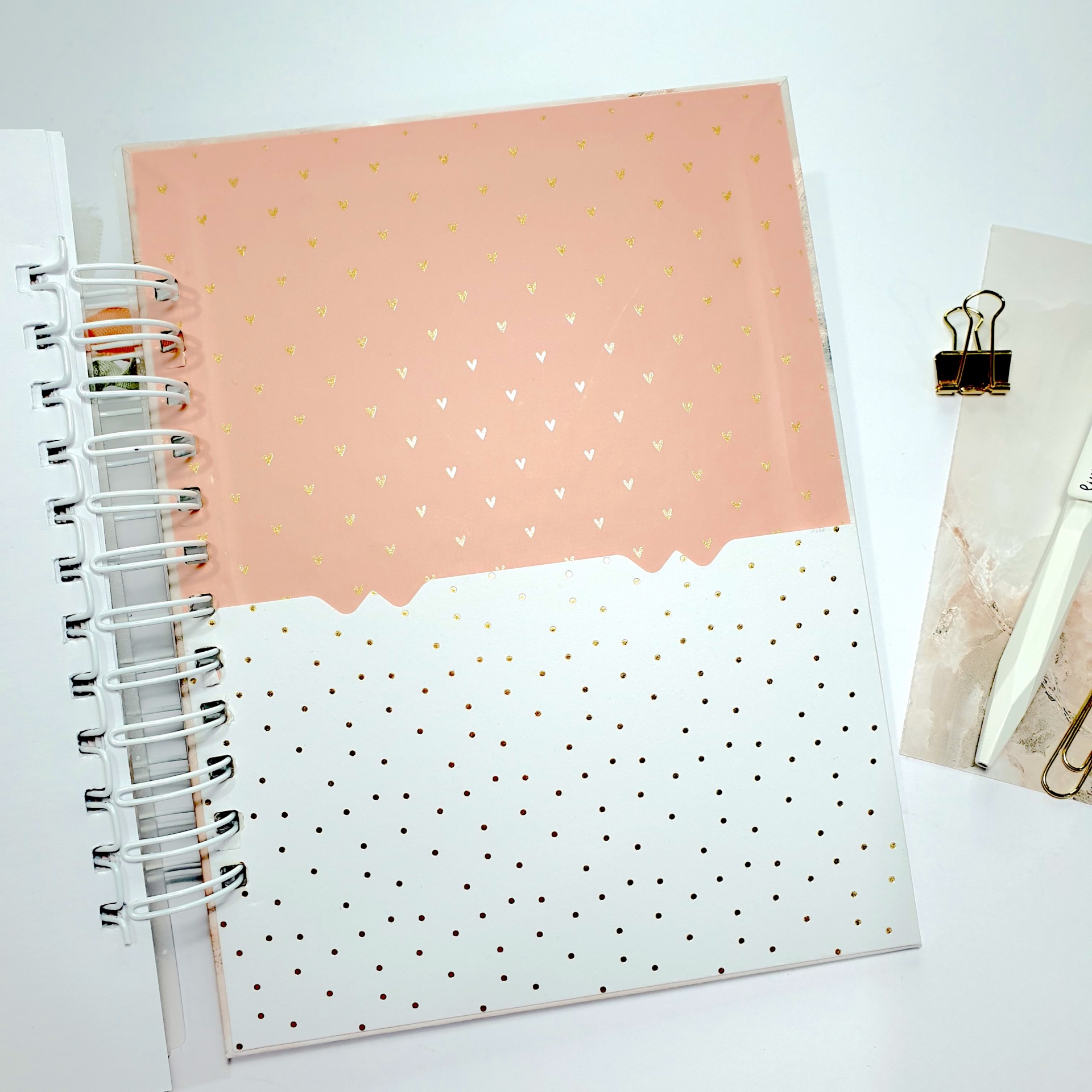 Caderno espiral rosa e branco com detalhes dourados, clipe preto e caneta branca sobre superfície branca