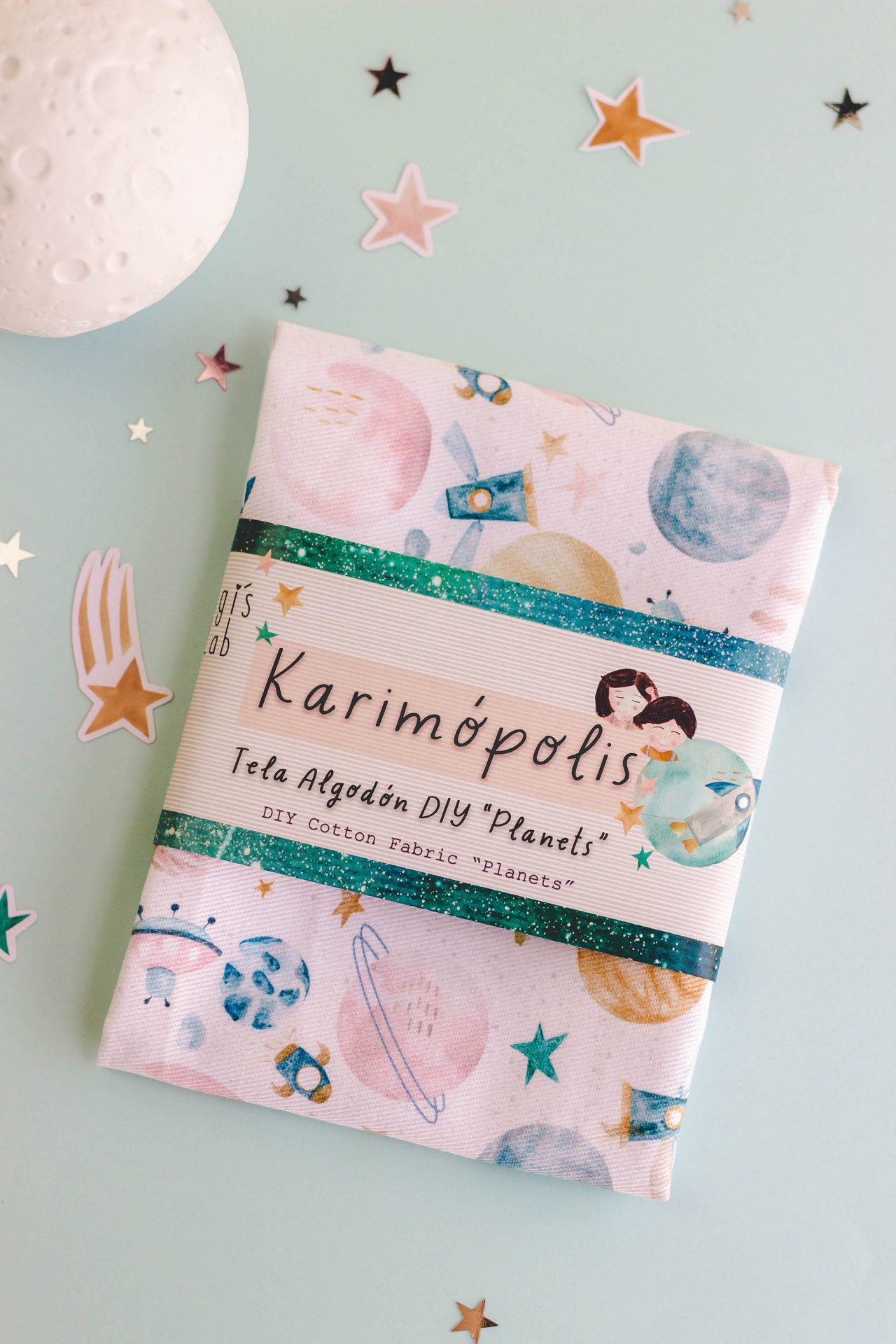 Tecido de algodão estampado com planetas e estrelas em embalagem sobre mesa azul clara com decoração de estrelas e globo lunar.