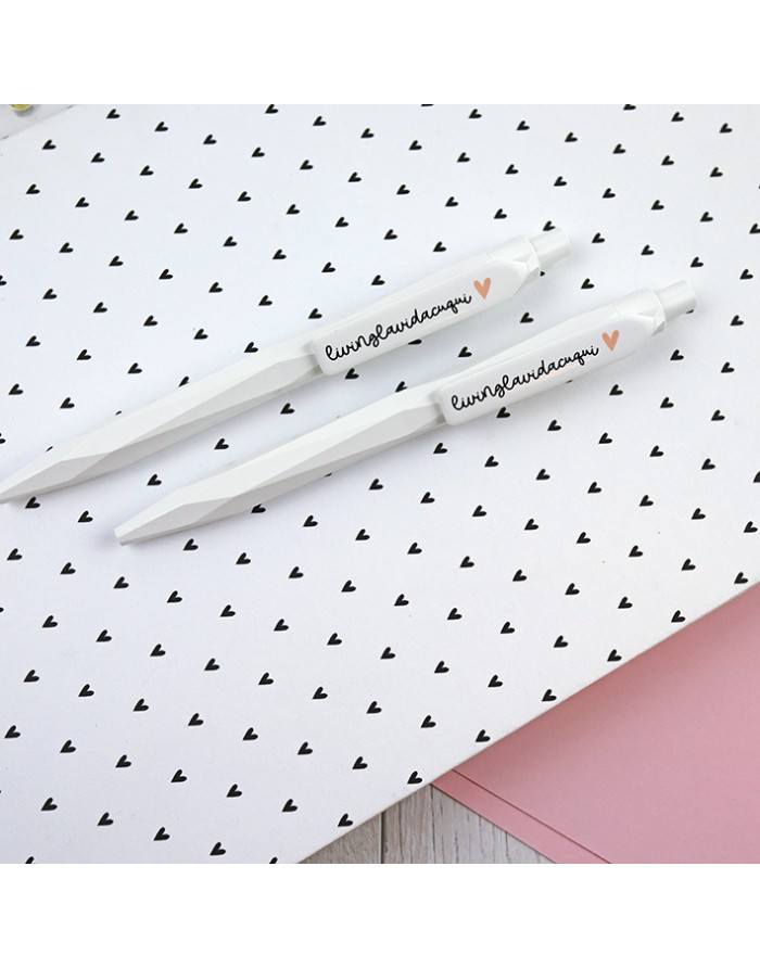 Dois esferográficos brancos com escrita e coração laranja sobre papel com corações pretos.