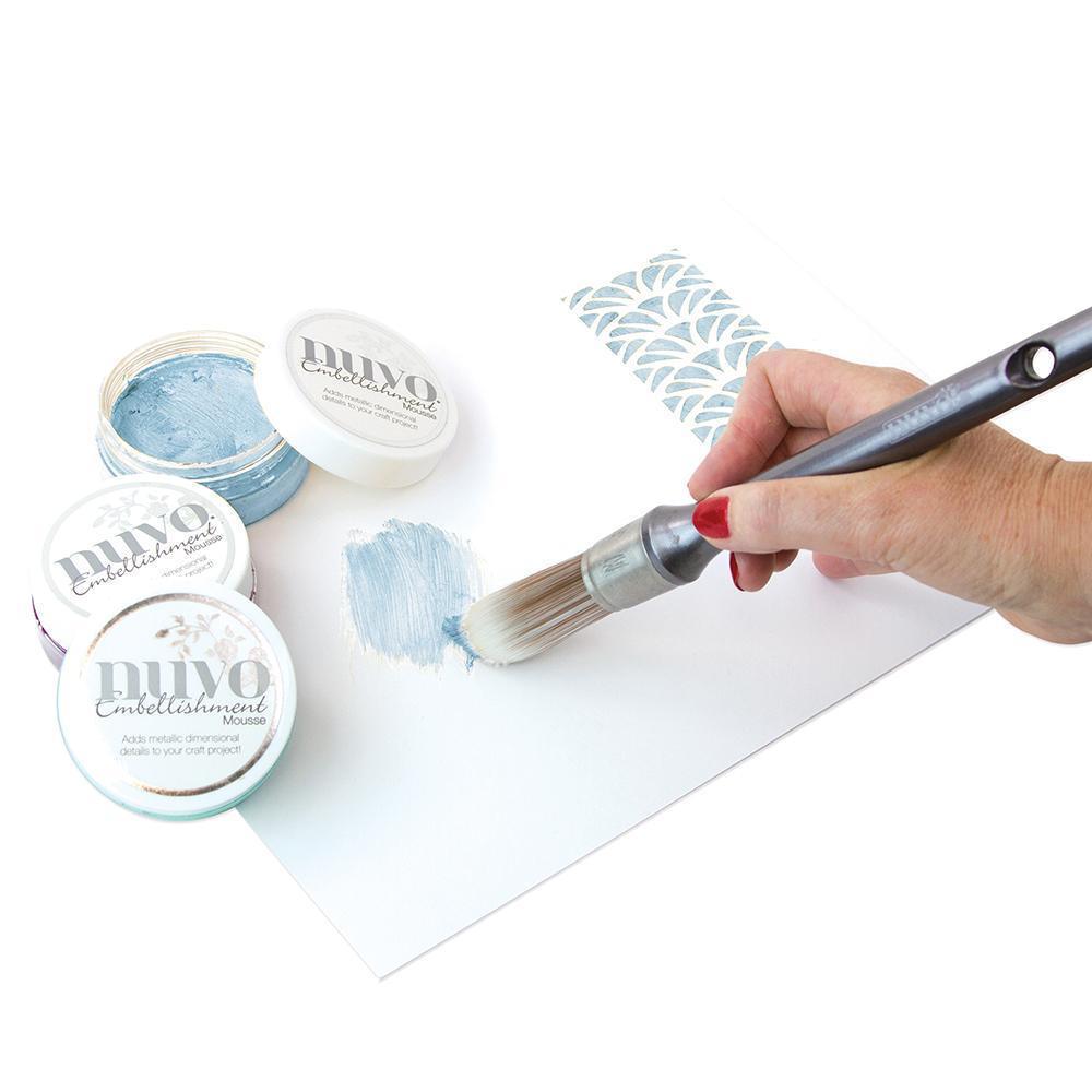 Pessoa a pintar com pincel tinta azul clara em papel branco ao lado de frascos de tinta