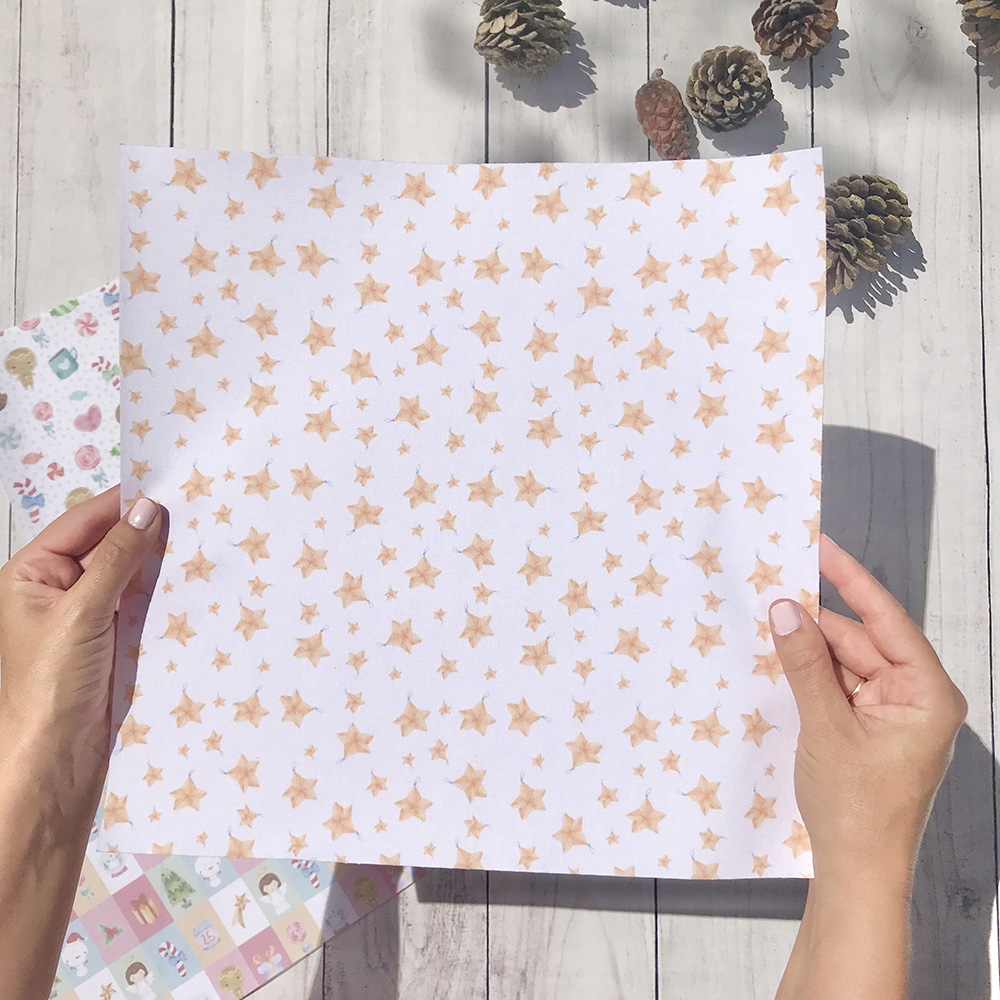 Pano branco com estrelas cor de rosa e laranja segurado por duas mãos sobre mesa de madeira clara com pinhas e papéis coloridos